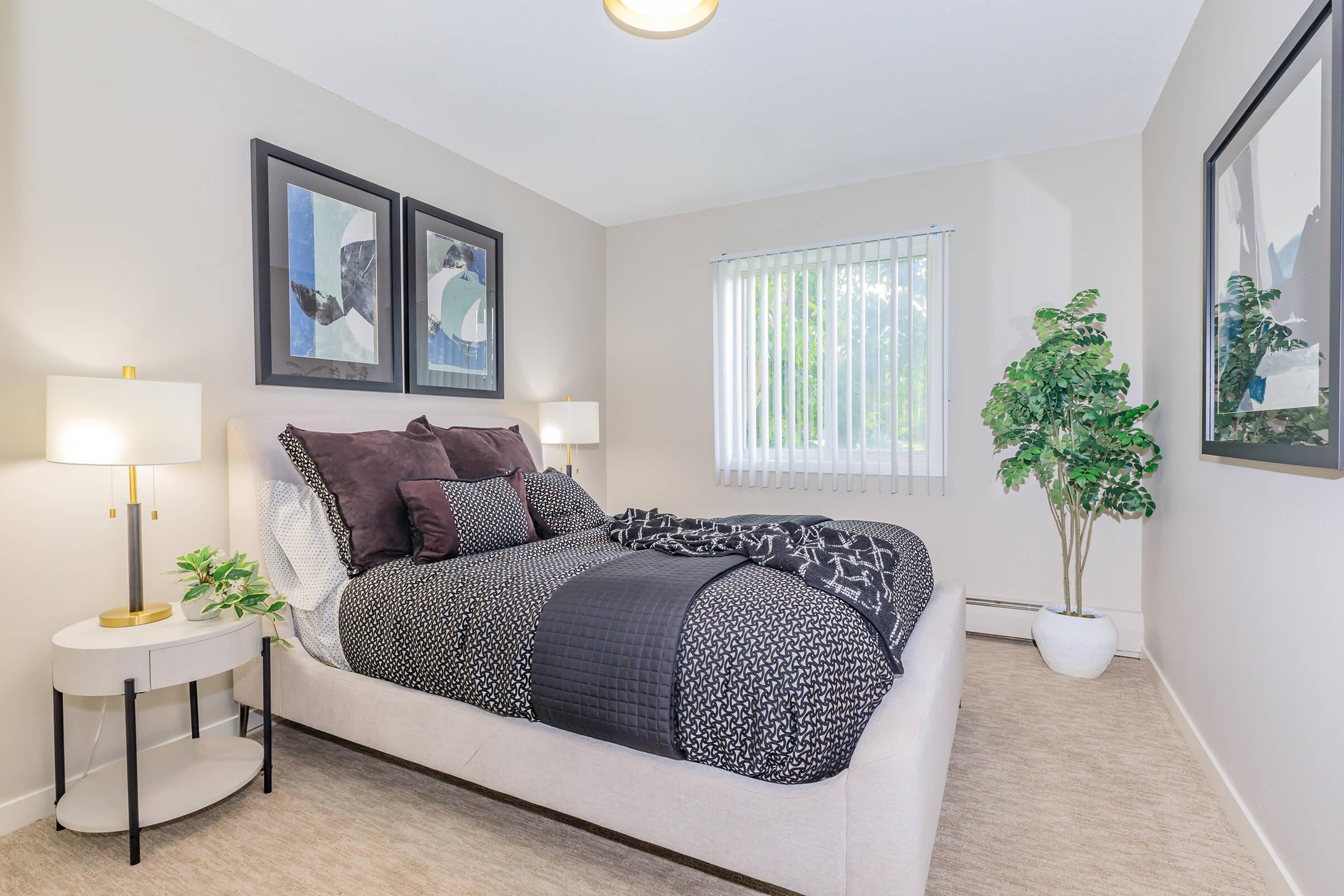 A cozy bedroom featuring a neatly made bed with dark bedding, two framed artworks on the wall, a small side table with a lamp, and a potted plant. Large windows with vertical blinds allow natural light to illuminate the space, creating a warm and inviting atmosphere.