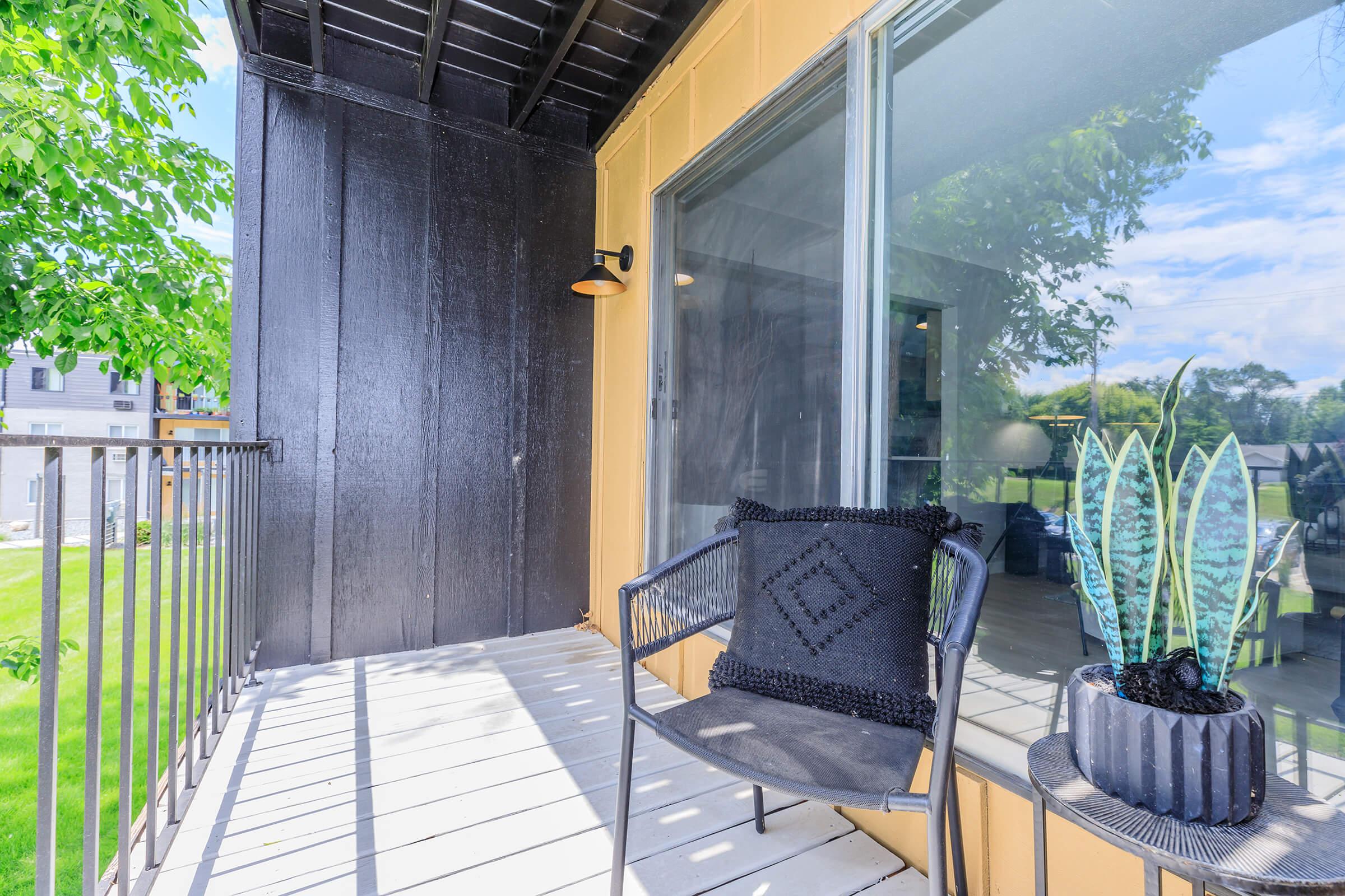 A balcony featuring a black chair with a textured cushion, a potted plant, and a modern wall light. The back wall is painted black, while the floor and railing are white. Green foliage can be seen outside, adding a touch of nature to the urban setting. A sunny sky is partially reflected in the glass door.