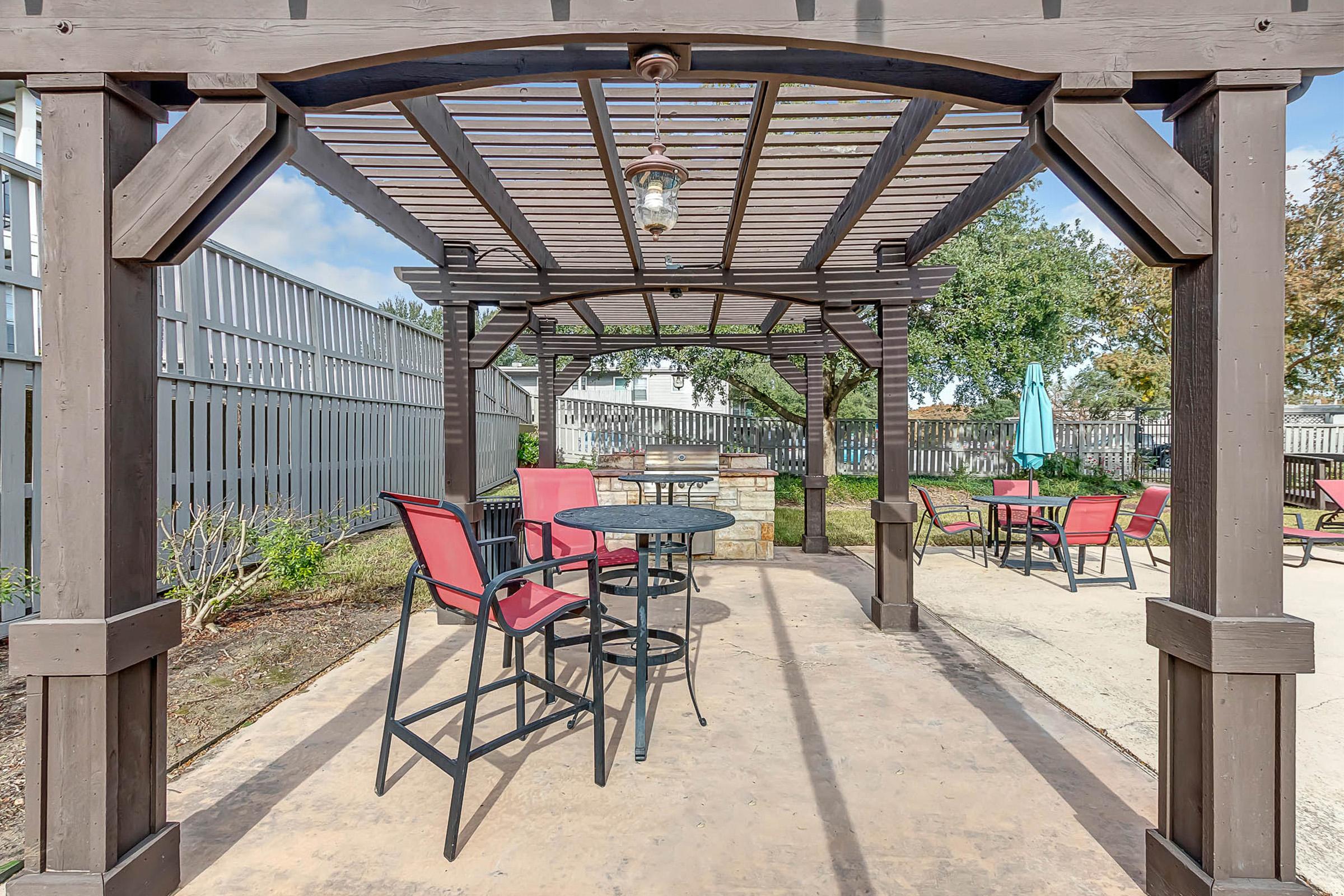 A shaded outdoor patio area with a wooden pergola, featuring a round black table and several red chairs. Surrounding the patio are green plants and a fence in the background, with an umbrella visible in the distance. The scene is well-lit with blue skies above.