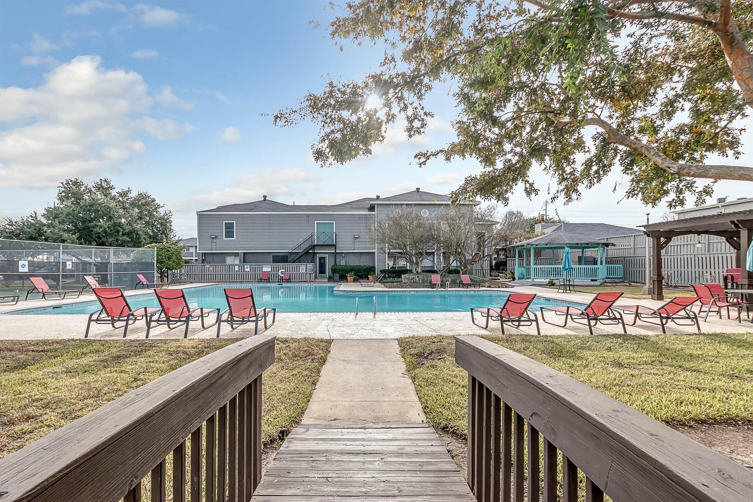 View of a swimming pool surrounded by lounge chairs, with a wooden walkway leading to the pool area. In the background, there are residential buildings and trees, under a partly cloudy sky. The scene evokes a relaxing outdoor atmosphere.