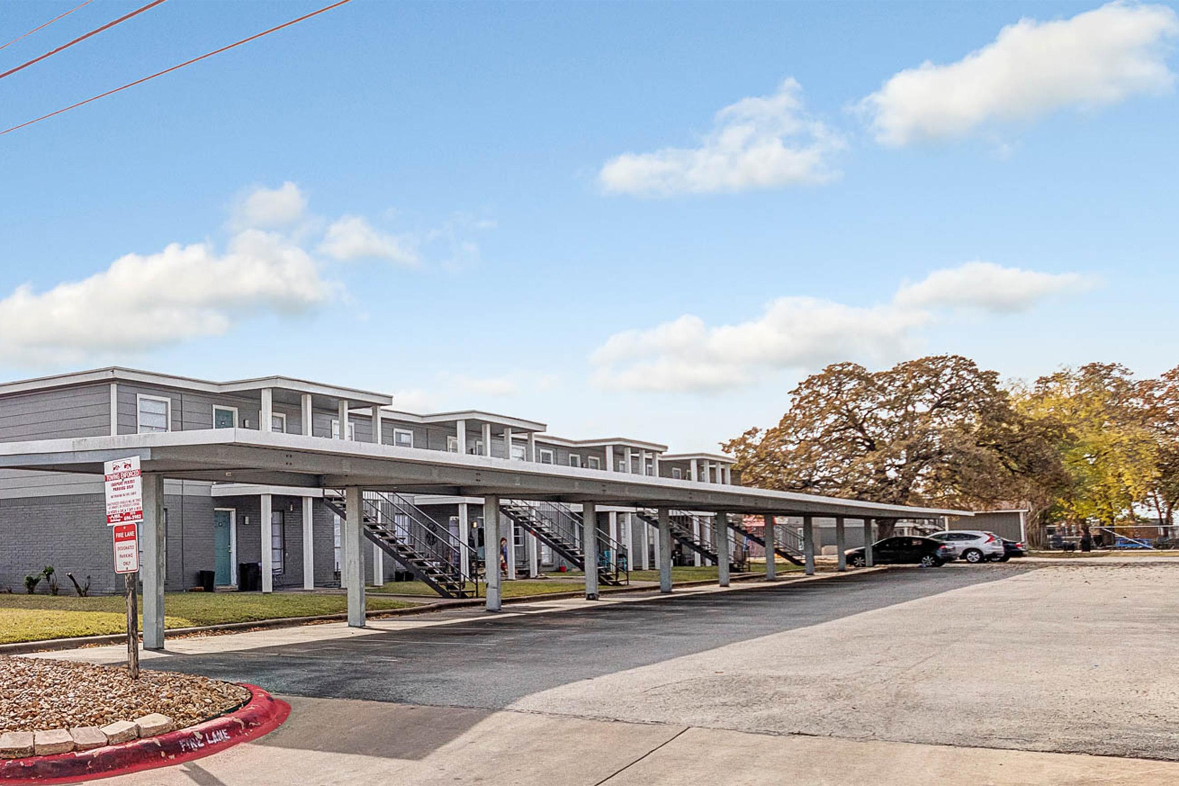 A row of modern, two-story apartment buildings with covered parking spaces underneath. The buildings have a light exterior and surrounded by grassy areas and trees. The sky is clear with a few clouds, and the scene conveys a suburban residential setting.