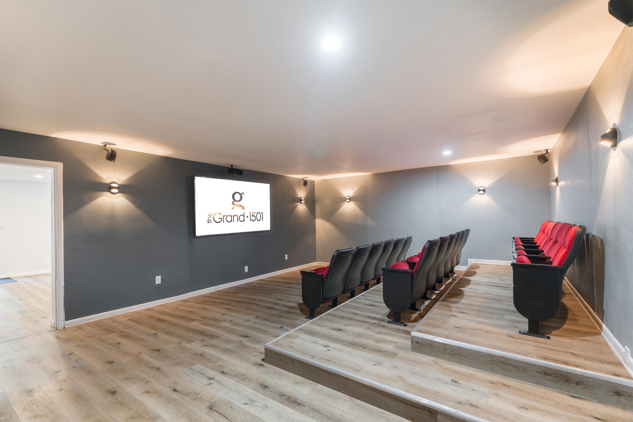 A modern home theater featuring tiered seating with black seats and red accents, a large screen displaying the name "The Grand 1501," gray walls, and stylish lighting fixtures. The floor is wooden, creating a comfortable and inviting atmosphere for movie watching.