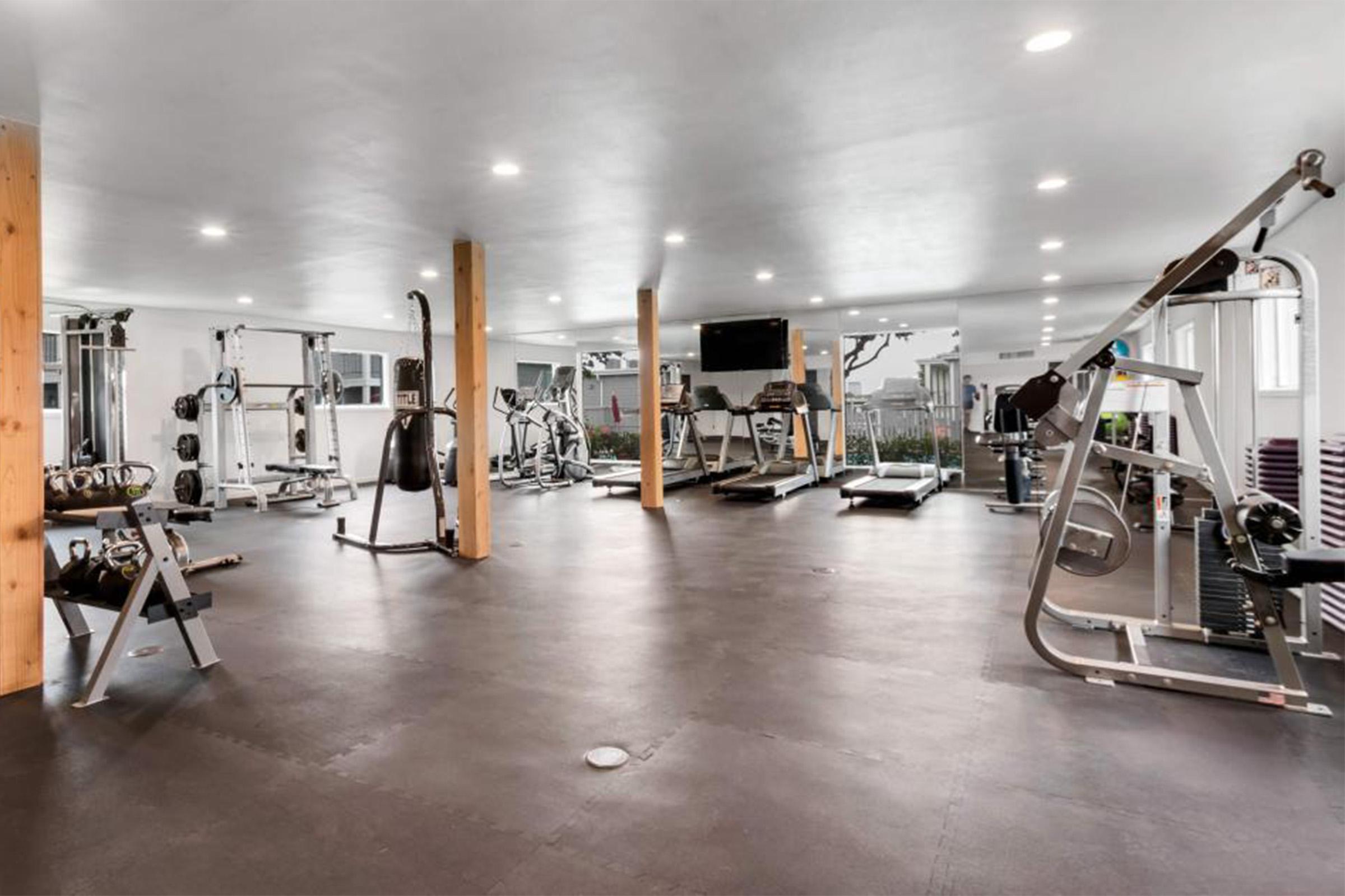 A spacious gym interior featuring various exercise equipment, including treadmills, weight machines, and free weights. The room is well-lit with overhead lighting, has wooden pillars, and offers a clean, modern design with a rubber floor. Large windows provide natural light.
