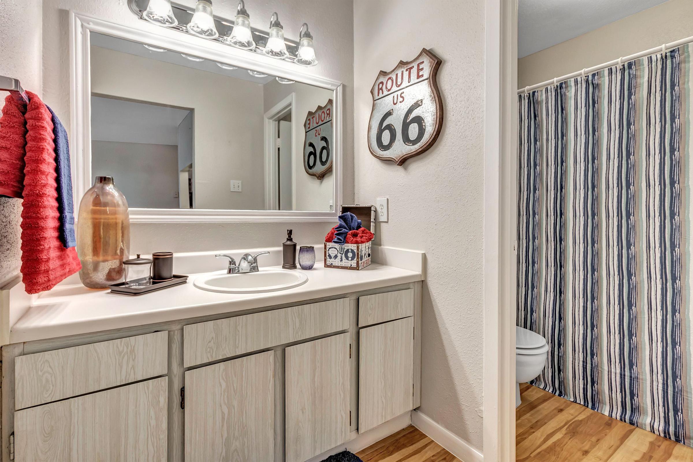 A bathroom with a large mirror above a countertop sink, featuring a striped shower curtain and Route 66 wall decor. There are various bath accessories, including a bottle, towels, and toiletries on the counter, with wooden flooring visible.