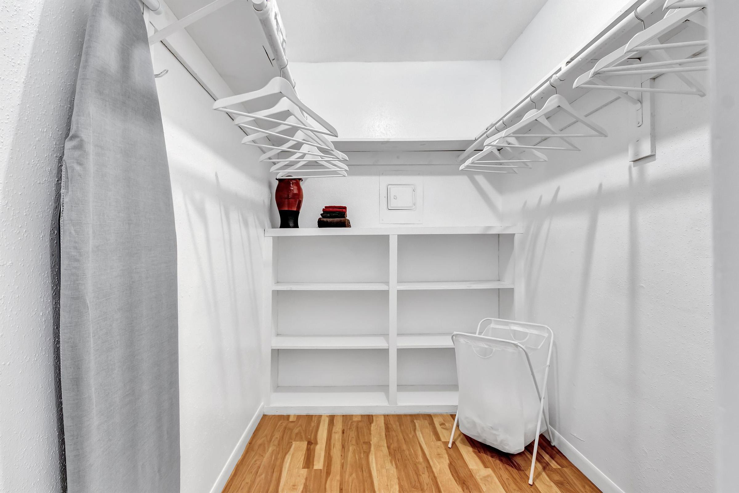 A spacious, empty closet with white walls and wooden flooring. It features multiple hanging rods, an open shelf unit, and a lightweight laundry basket. A gray curtain is draped on one side, and a few folded towels or clothes are neatly placed on the top shelf. The overall ambiance is clean and uncluttered.