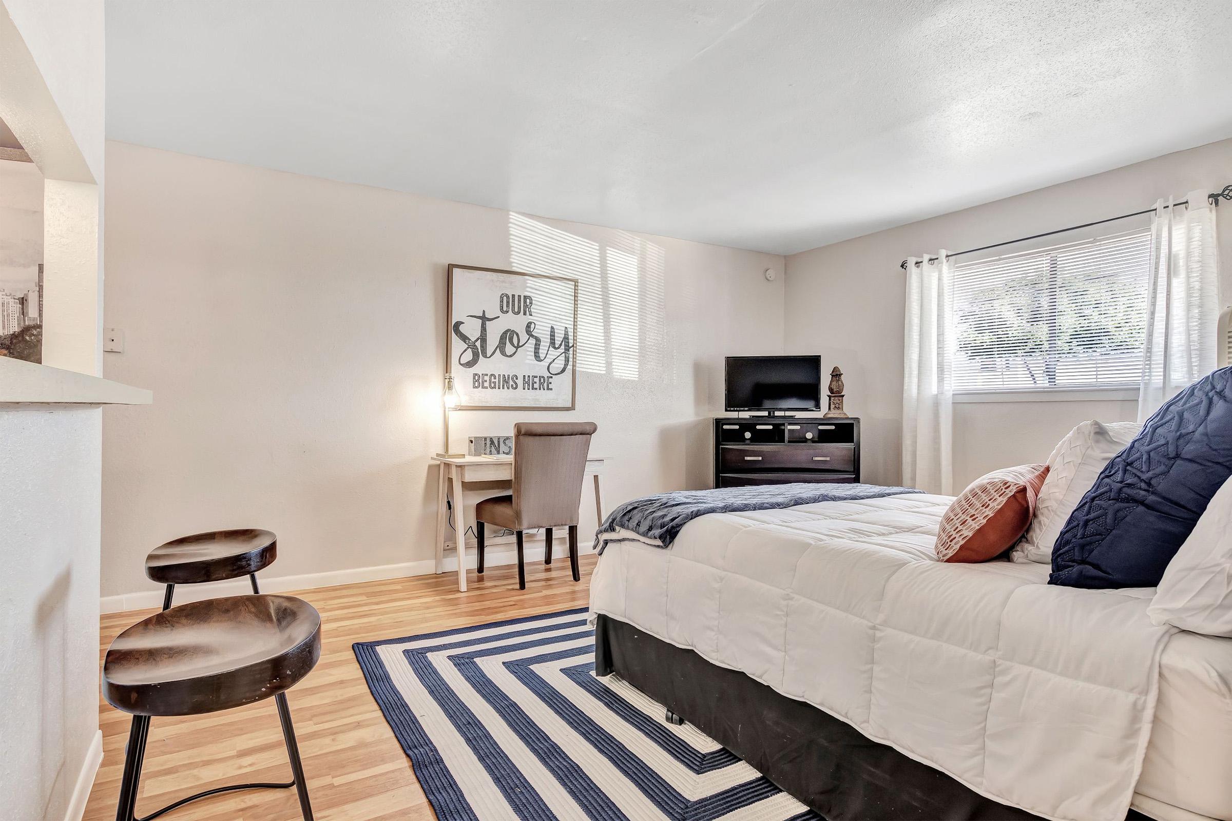 A cozy bedroom featuring a queen-sized bed with white bedding, a navy blue throw blanket, and decorative pillows. There is a small desk and chair against one wall, a dresser, and a TV. Natural light filters through the window, and the wooden floor is complemented by a striped area rug. A wall art piece reads "Our Story Begins Here."