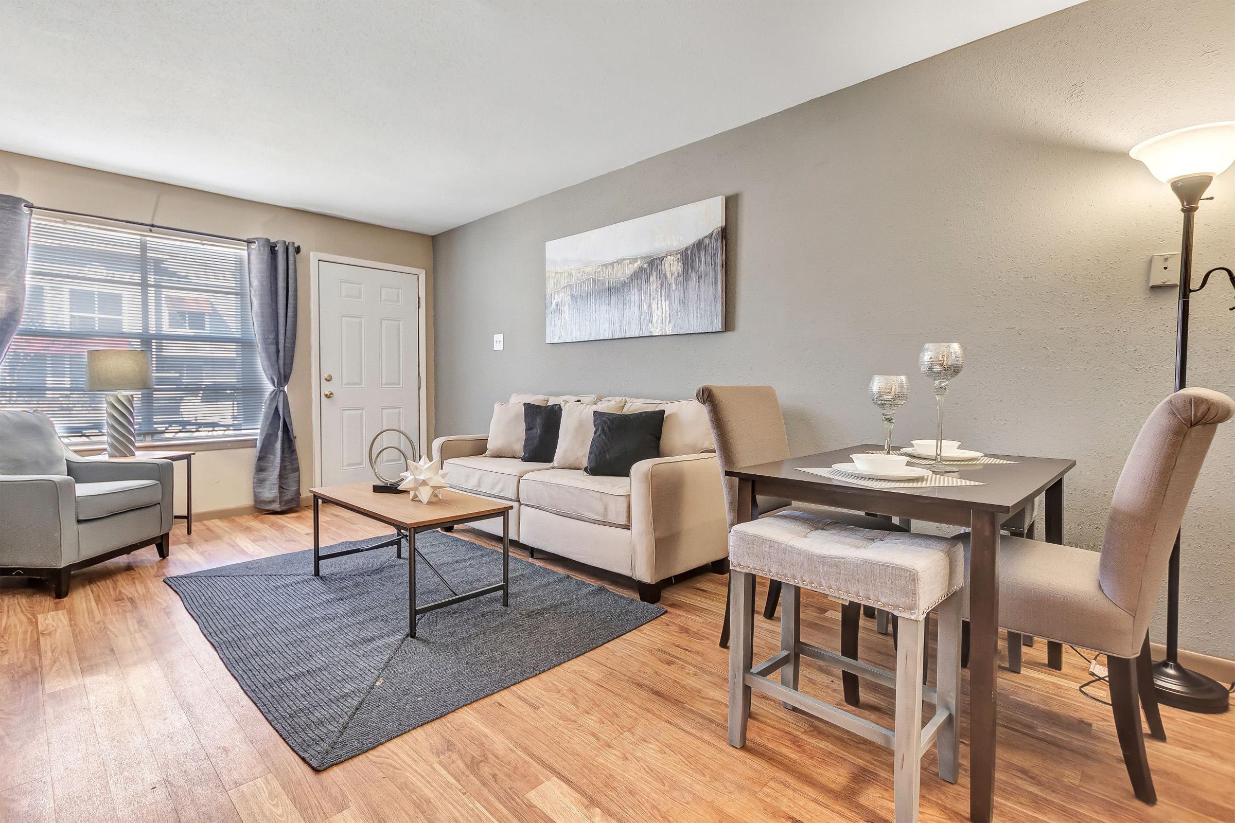 A cozy living room with a light gray couch, two black cushions, and a small coffee table. A dining table with two chairs and two place settings is nearby. A large window lets in natural light, and there’s a lamp illuminating the space. The floor is wood, and a textured area rug complements the decor.