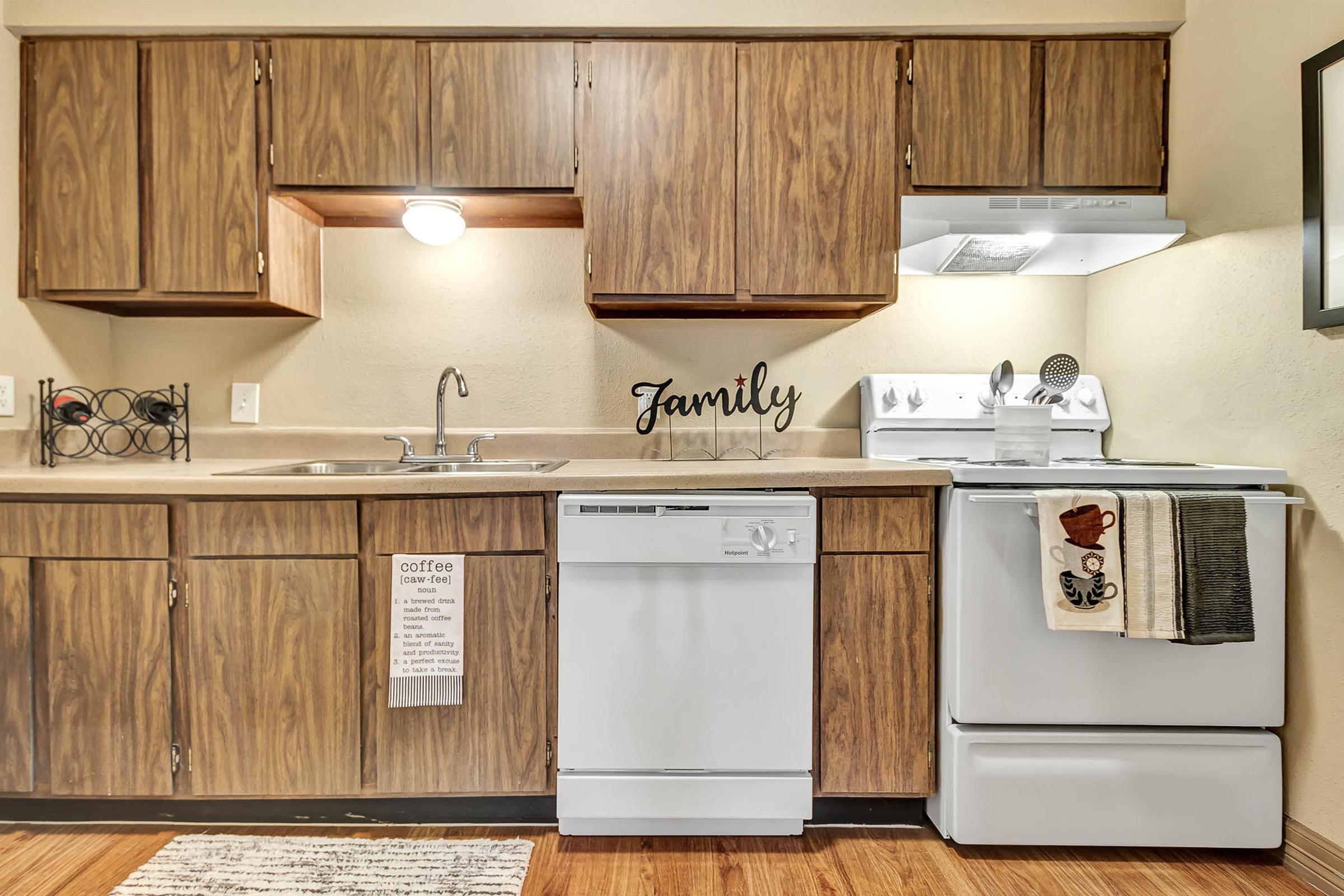 A warm and inviting kitchen featuring wooden cabinets, a stainless steel sink, a dishwasher, and a white stove. A decorative "Family" wall hanging adds a personal touch. A dish towel with a coffee theme hangs beside the sink, and a textured rug is placed on the wooden floor.