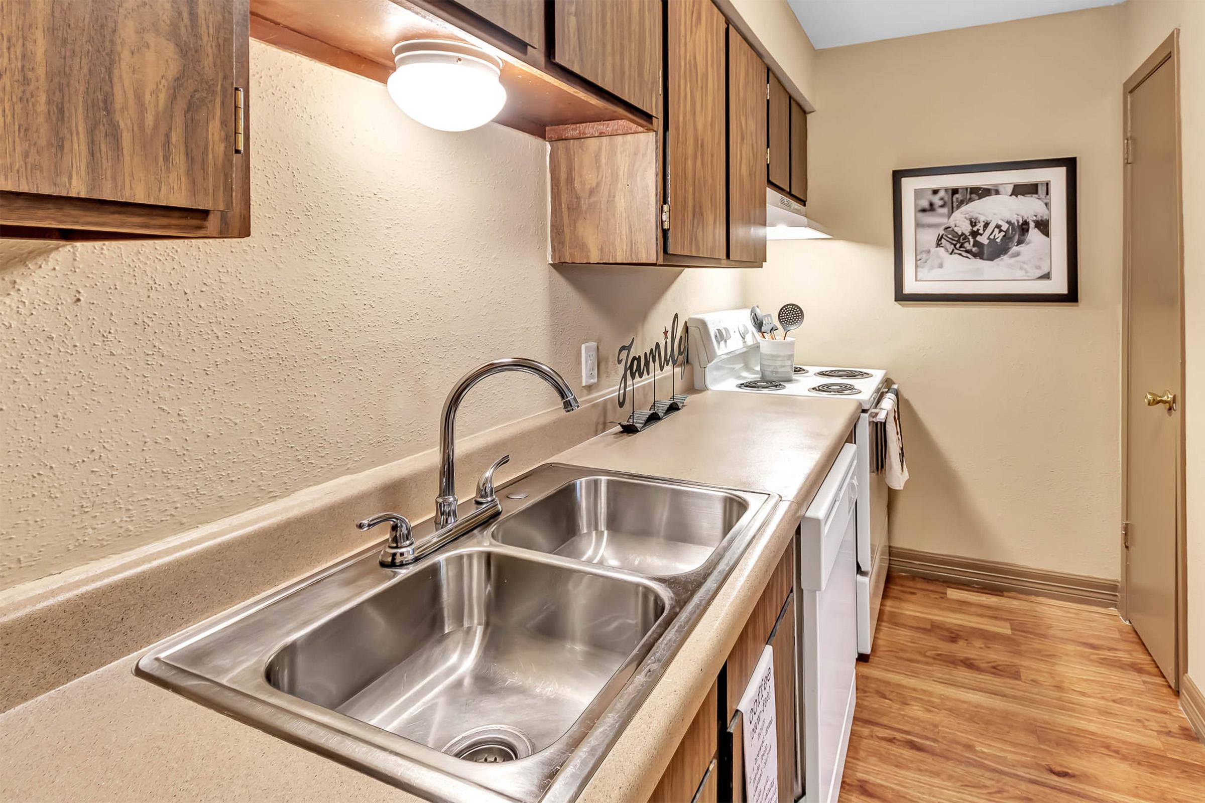 A modern kitchen featuring a light-colored countertop with double stainless steel sinks. Above the sink, there are wooden cabinets and a light fixture. The kitchen includes a stove and a framed picture on the wall, creating a warm and inviting atmosphere. The flooring is made of wood.