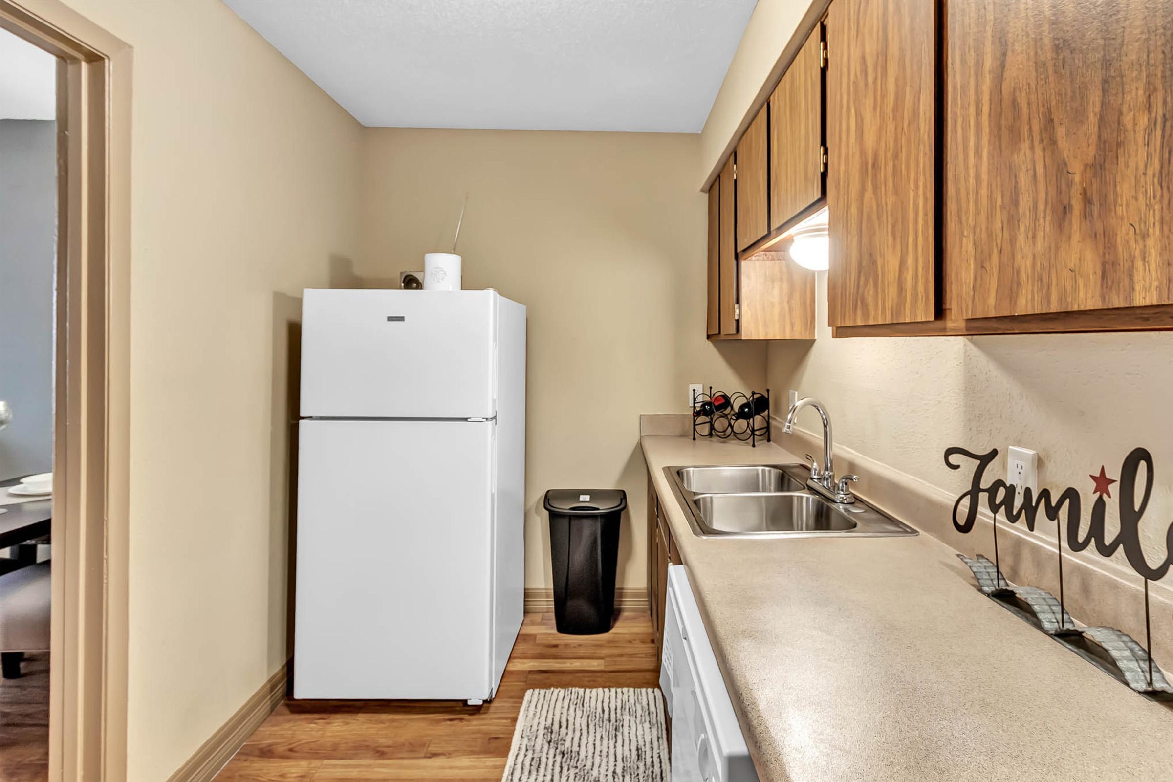 A cozy kitchen featuring a white refrigerator, sink, and cabinets with wooden doors. There is a trash can and decorative items on the countertop, including a sign that says "Family." The floor is wooden, and the walls are painted in a warm beige color, creating a welcoming atmosphere.