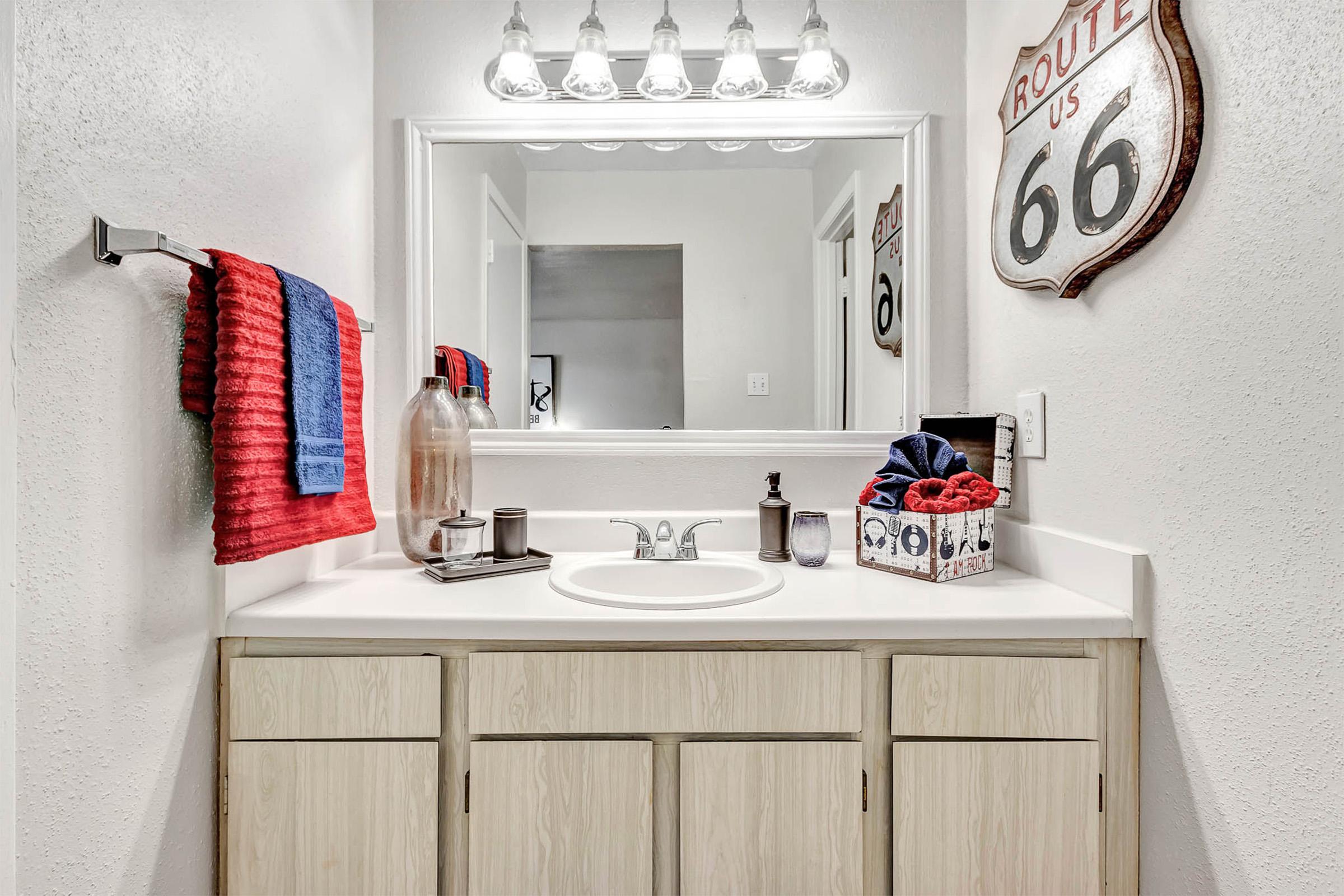 Bathroom scene featuring a white countertop with a sink, decorated with a soap dispenser, towels in red and blue, and a decorative container. Above the sink is a large mirror reflecting the space. A vintage Route 66 sign is mounted on the wall, adding a nostalgic touch to the decor.