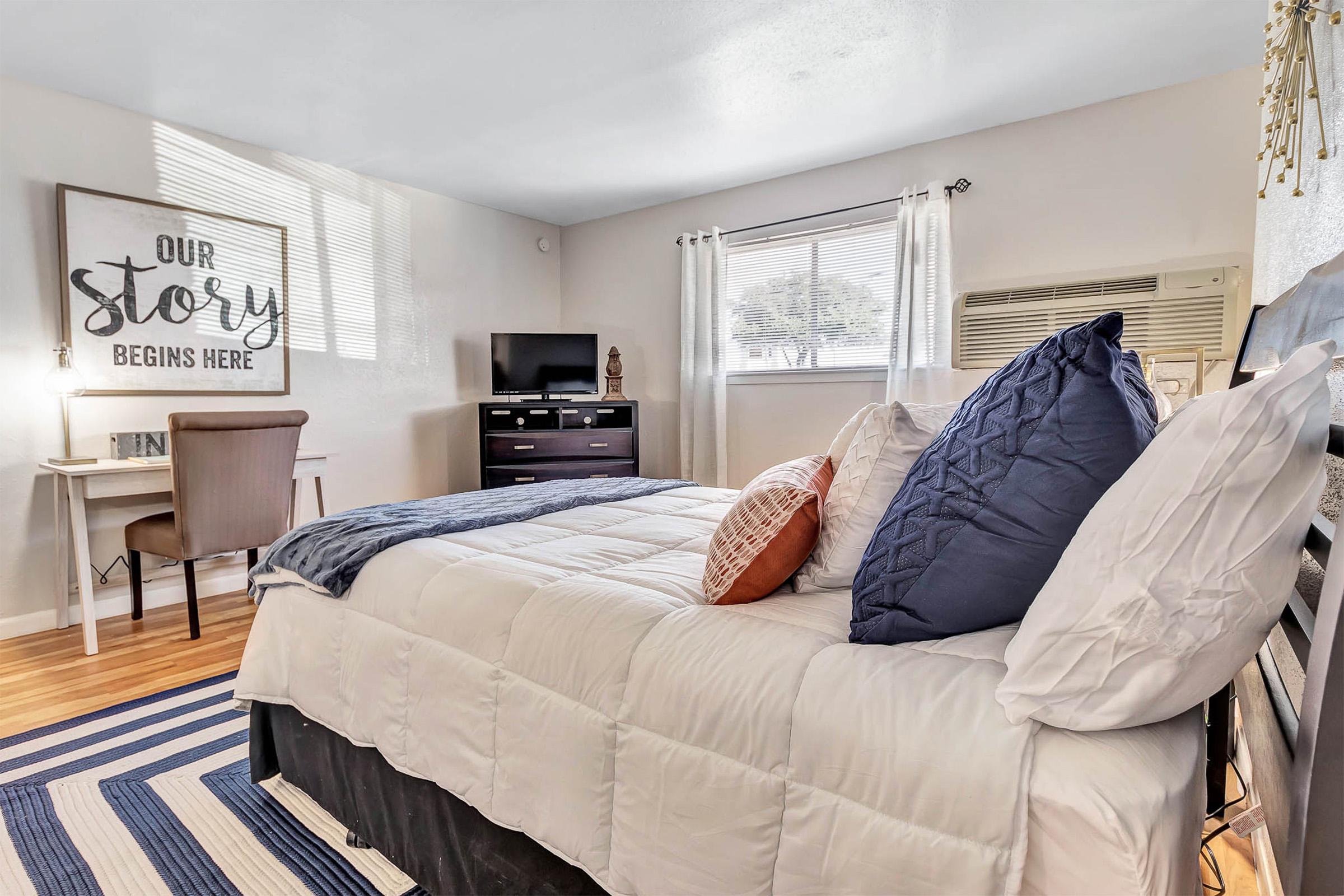 Cozy bedroom featuring a comfortable bed with white bedding and blue accent pillows, a decorative orange pillow, a desk with a chair, a flatscreen TV on a dresser, and a large window with sheer curtains. The walls are adorned with a framed sign that reads "Our Story Begins Here," adding a personal touch.