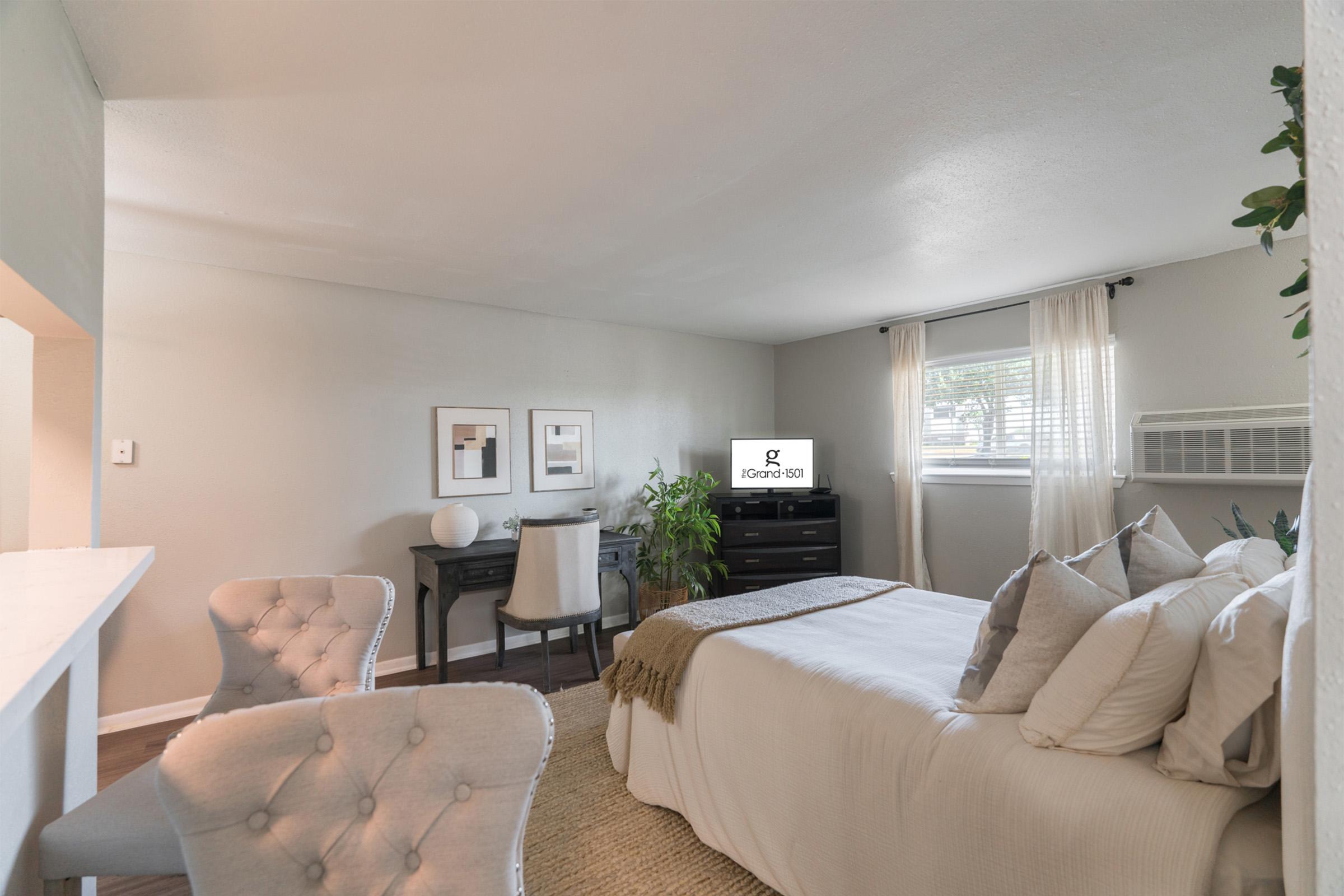 A cozy bedroom with a bed adorned with decorative pillows, two elegant chairs at the foot, a small desk with a chair, and a television on a dresser. The room has light-colored walls, a window with sheer curtains, and a green plant for a touch of nature.
