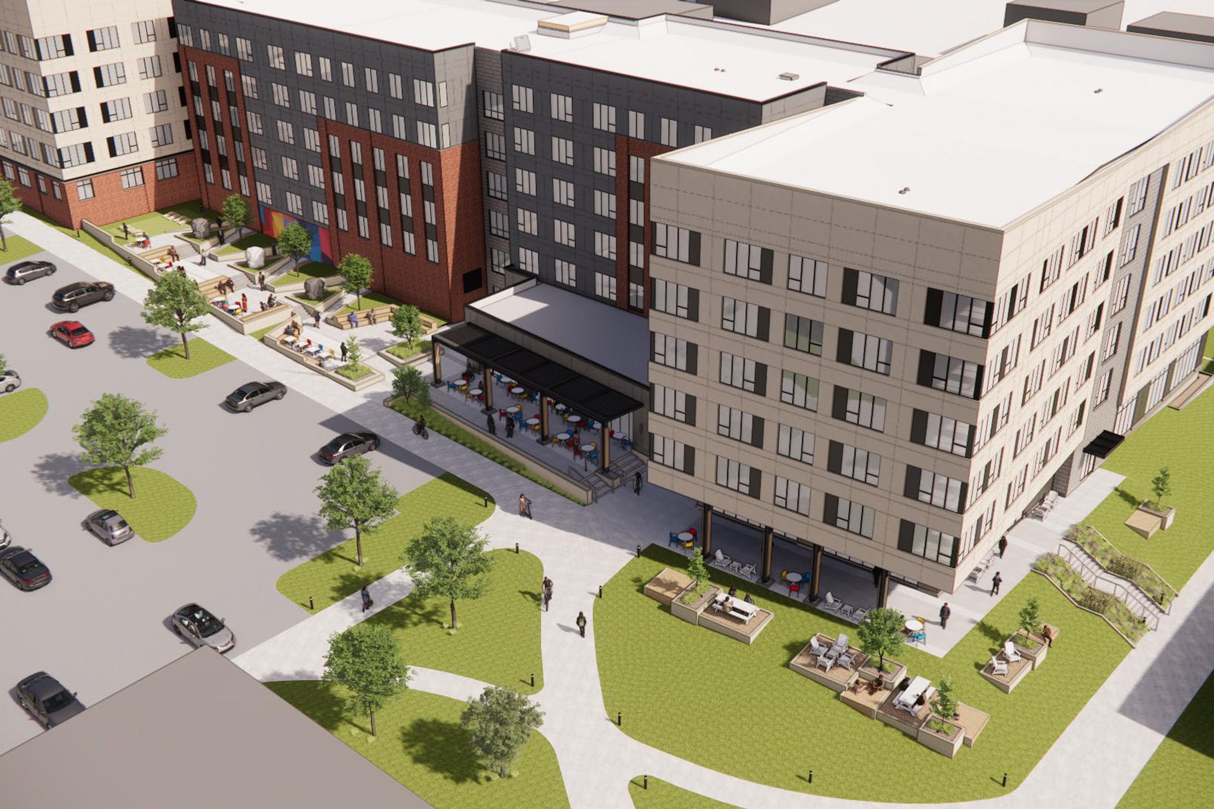 Aerial view of a modern residential complex featuring multiple buildings with a mix of brick and light-colored facades. There are landscaped areas with greenery, benches, and outdoor seating. Parked cars line the streets around the complex, and people can be seen walking and enjoying the outdoor spaces.