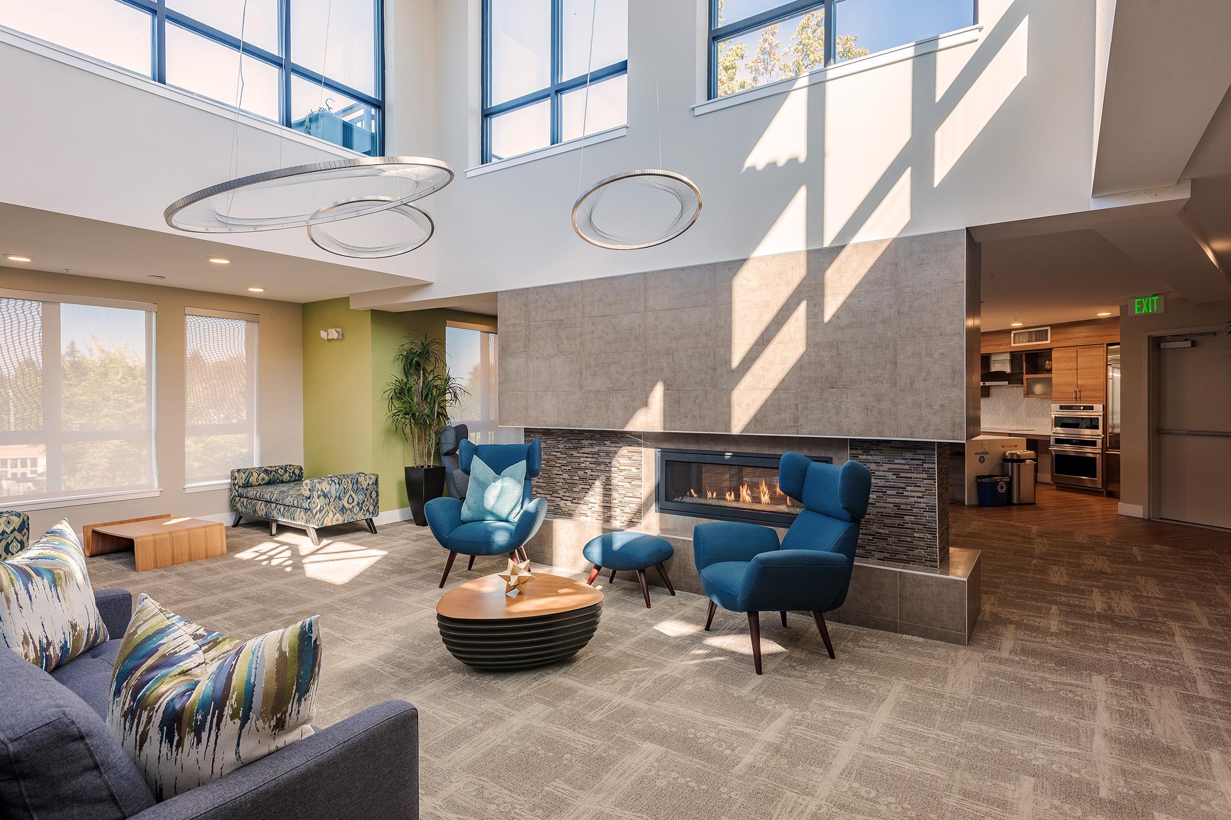 Modern lounge space with large windows allowing natural light. Features include two blue armchairs, a round coffee table, a gray sofa with patterned cushions, and a stone fireplace. The decor is contemporary with geometric light fixtures, and there are cozy seating areas and warm tones throughout the room.