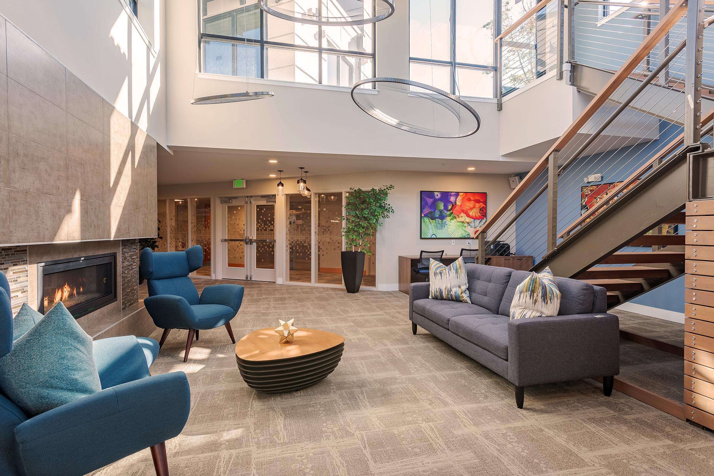 A modern, well-lit interior of a lounge area featuring a blue sofa, two blue accent chairs, a circular wooden coffee table, and a decorative fireplace. Large windows provide ample natural light, and artwork adorns the walls. A staircase with wooden railings leads to an upper level.