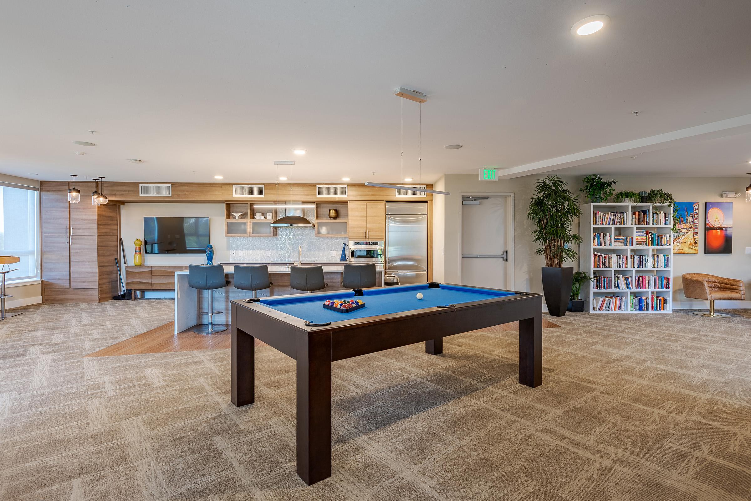 A spacious modern lounge area featuring a blue billiard table at the center. In the background, there is a stylish kitchen with bar stools, a flat-screen TV, and a bookshelf filled with books. Large windows allow natural light to fill the room, creating a welcoming atmosphere.