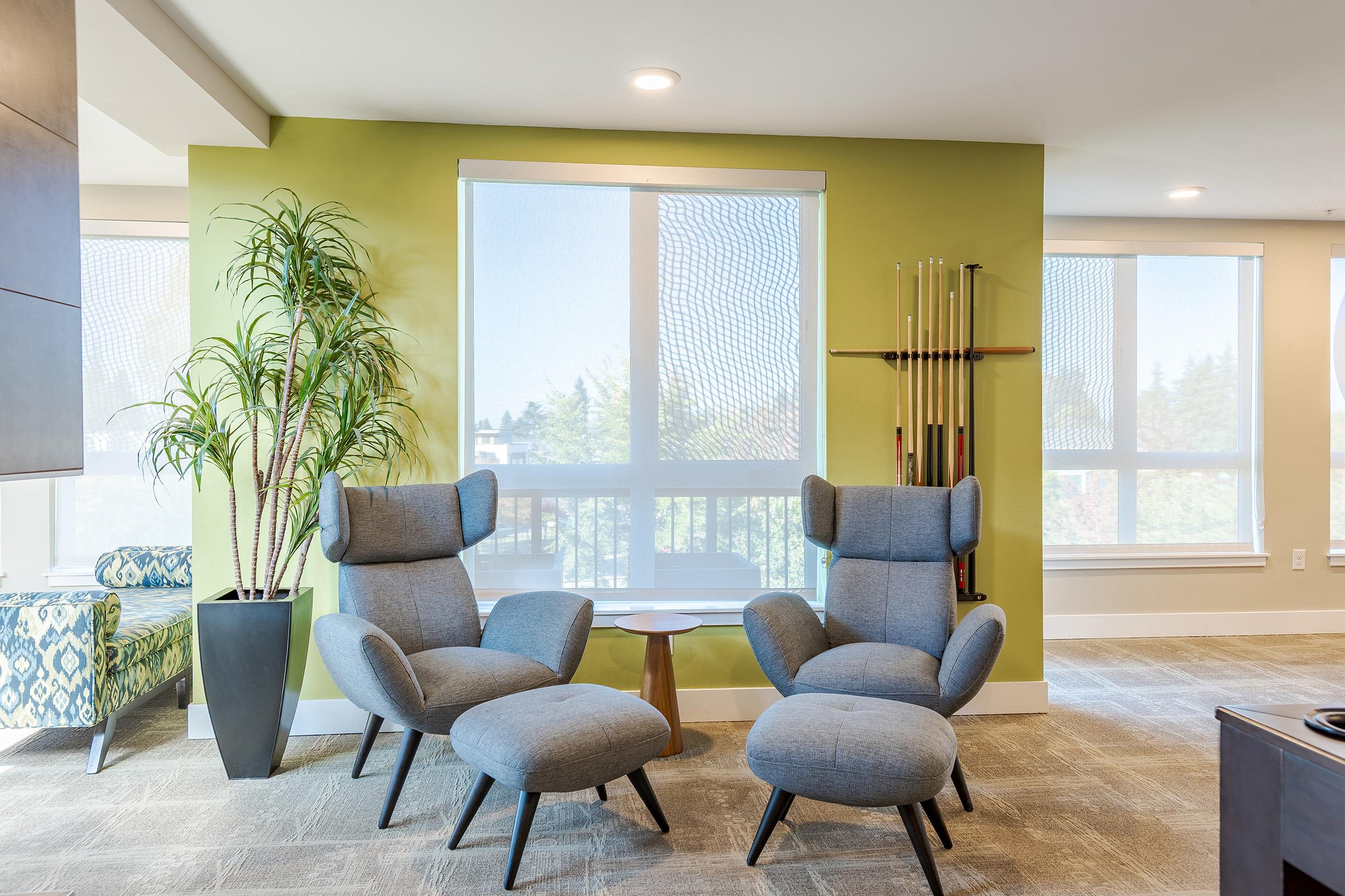 A cozy lounge area featuring two modern gray chairs with matching ottomans, a small round wooden side table, and a tall indoor plant in a black planter. Light filters through large windows with sheer blinds, illuminating the room with a fresh, inviting atmosphere. A wall-mounted rack with pool cues adds a recreational touch.