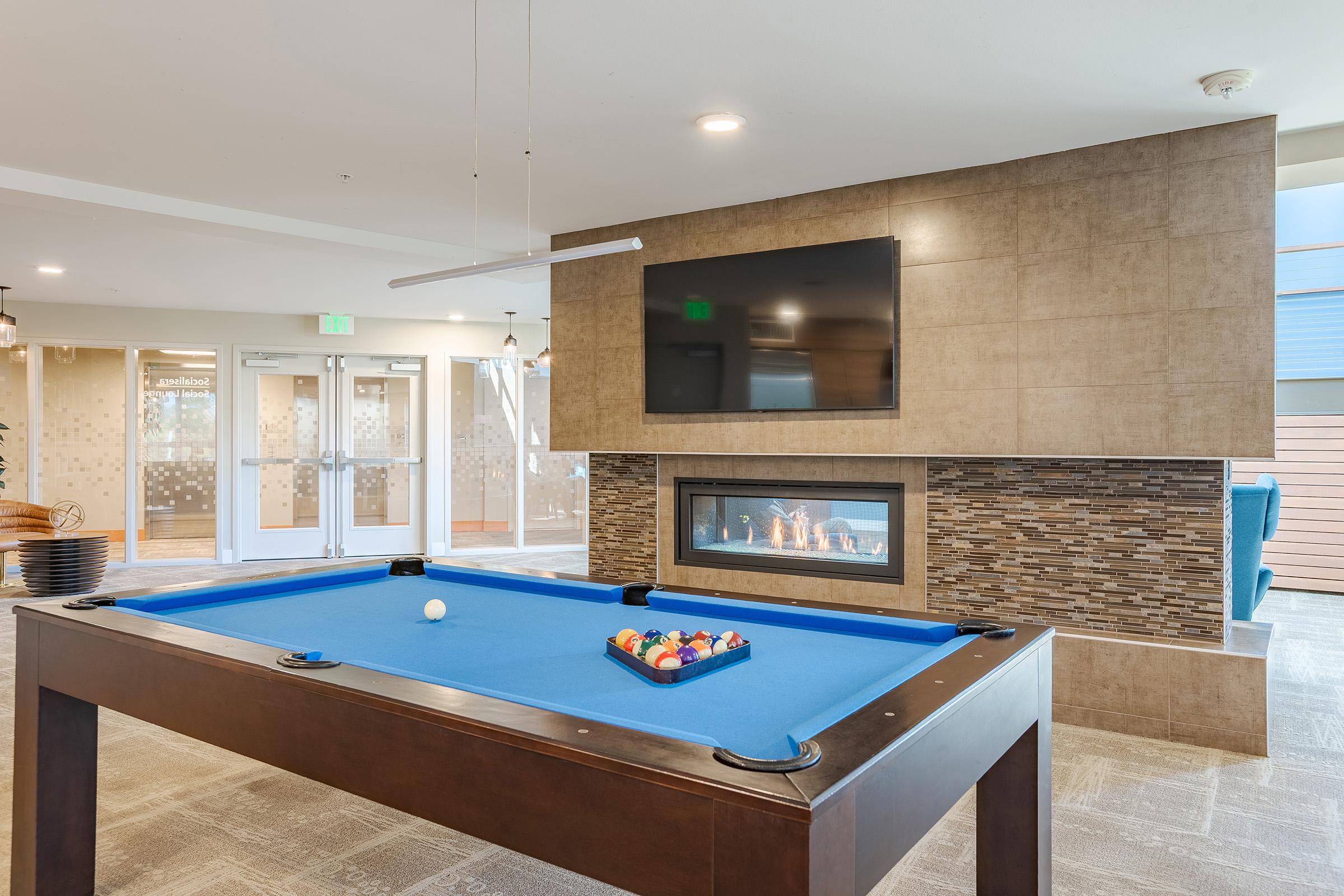 A modern game room featuring a blue pool table with a cue ball and a rack of colorful pool balls. In the background, there is a flat-screen TV mounted on a stylish wall with a cozy fireplace beneath it. The space has a contemporary design with neutral-colored walls and ambient lighting.
