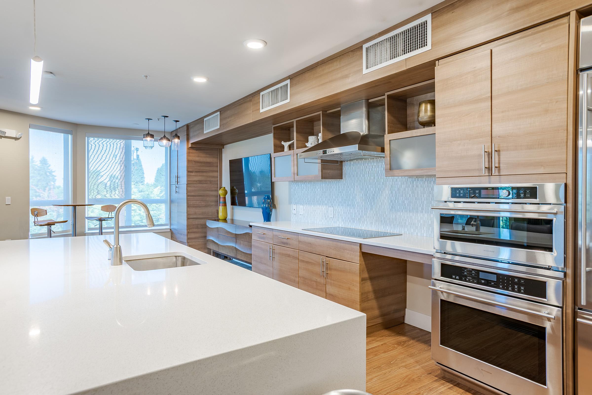 Modern kitchen with wooden cabinetry, a large white countertop, and stainless steel appliances. Features include a double wall oven, a sleek range hood, and an open space with a dining area visible in the background. Natural light floods through large windows, enhancing the contemporary design.
