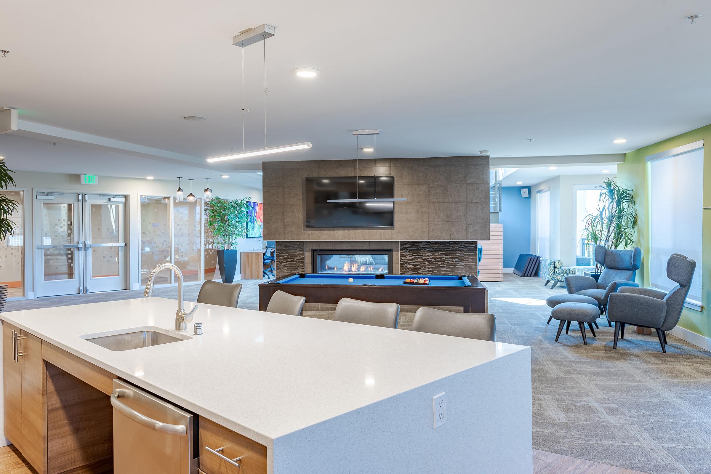 A modern communal space featuring a kitchen island with a sink, a cozy seating area with chairs, a fireplace, and a pool table. Natural light and greenery enhance the inviting atmosphere. The room is designed for relaxation and social gatherings.