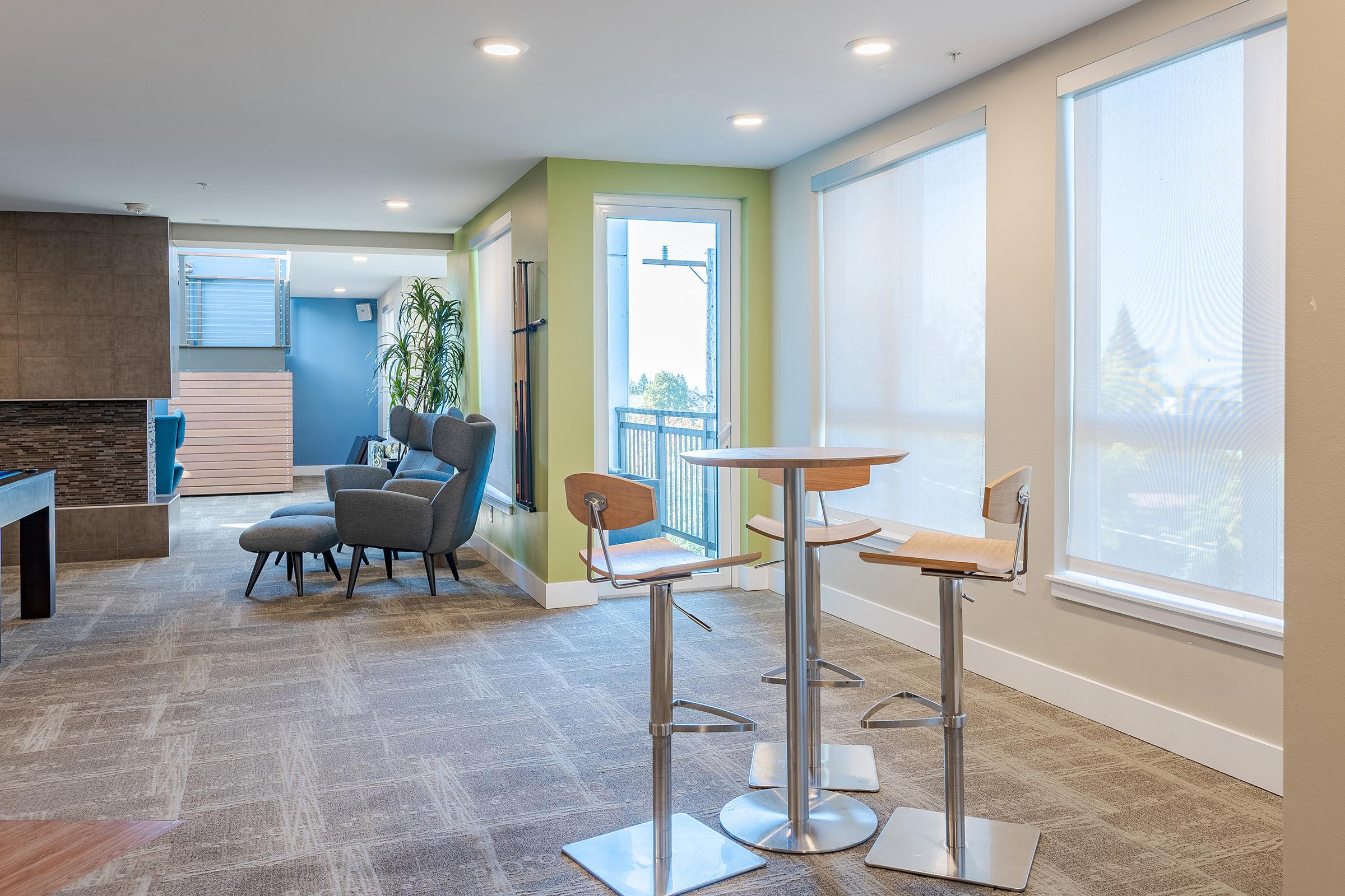 A modern interior space featuring a small round table with two tall stools, an accent chair, and large windows with sheer blinds. The walls are painted in a light green and grey color scheme, creating a bright and inviting atmosphere. Natural light fills the room, enhancing its contemporary design.