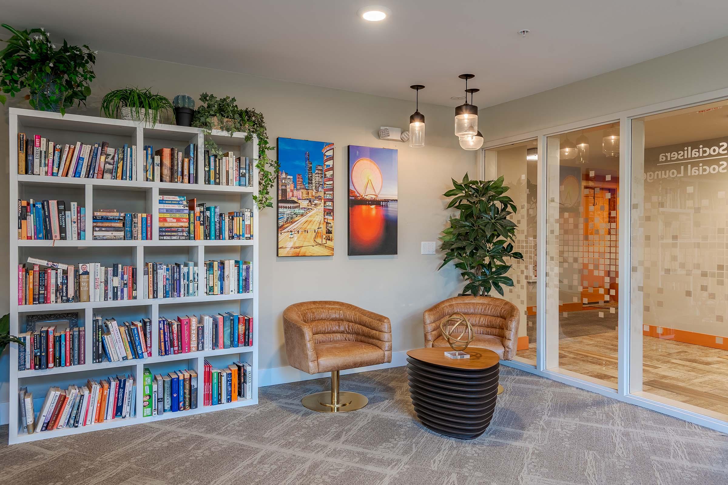A cozy reading nook featuring a large bookshelf filled with various books, two comfortable brown armchairs, a round wooden table, and decorative plants. In the background, there is a glass-walled meeting room with the words "social lounge" visible. The space is well-lit and inviting.