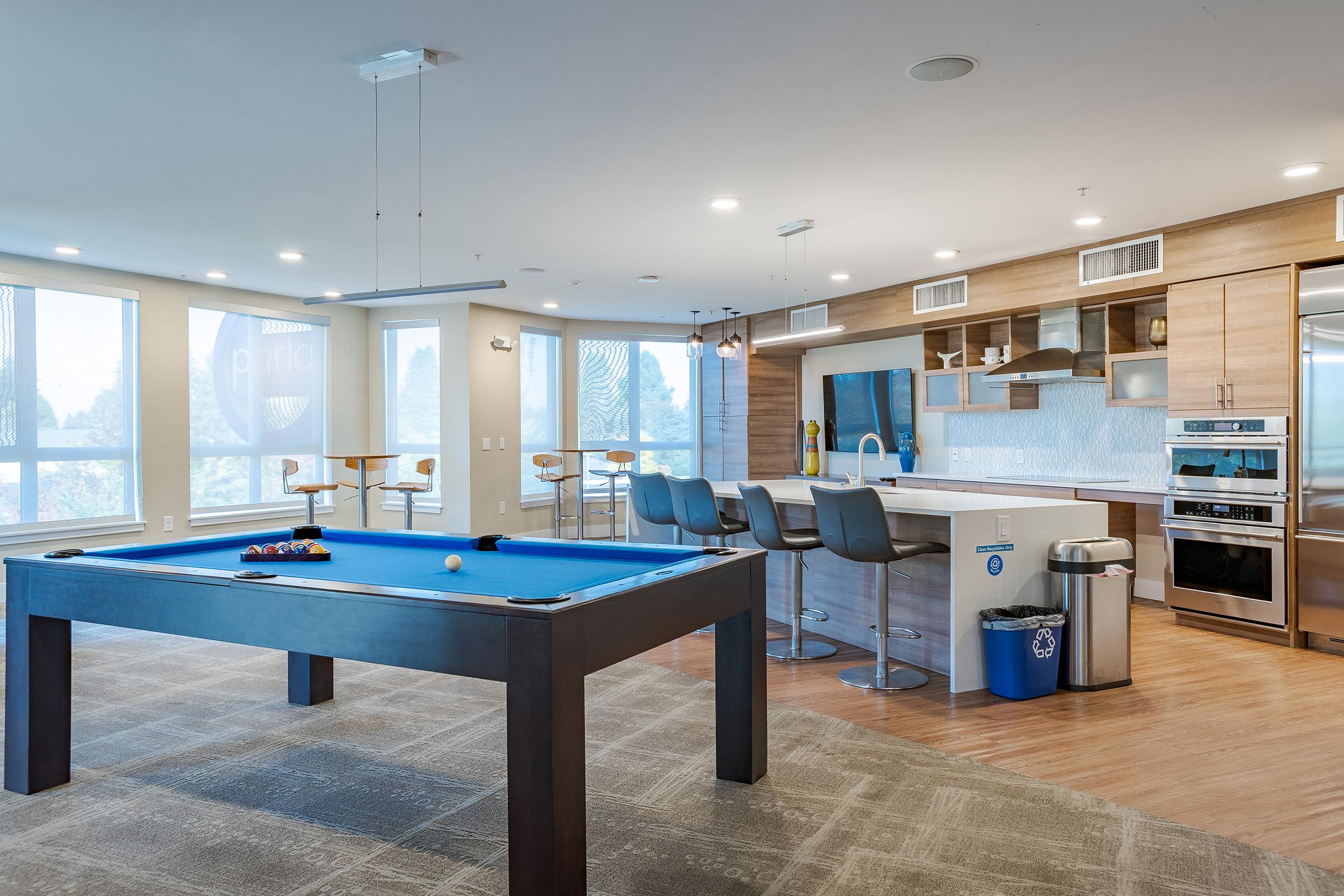 A modern recreational room featuring a blue pool table in the foreground. In the background, there are bar stools at a kitchen island with stylish cabinetry and appliances. Large windows allow natural light, and the space has an inviting, contemporary aesthetic. A trash bin is visible near the kitchen area.
