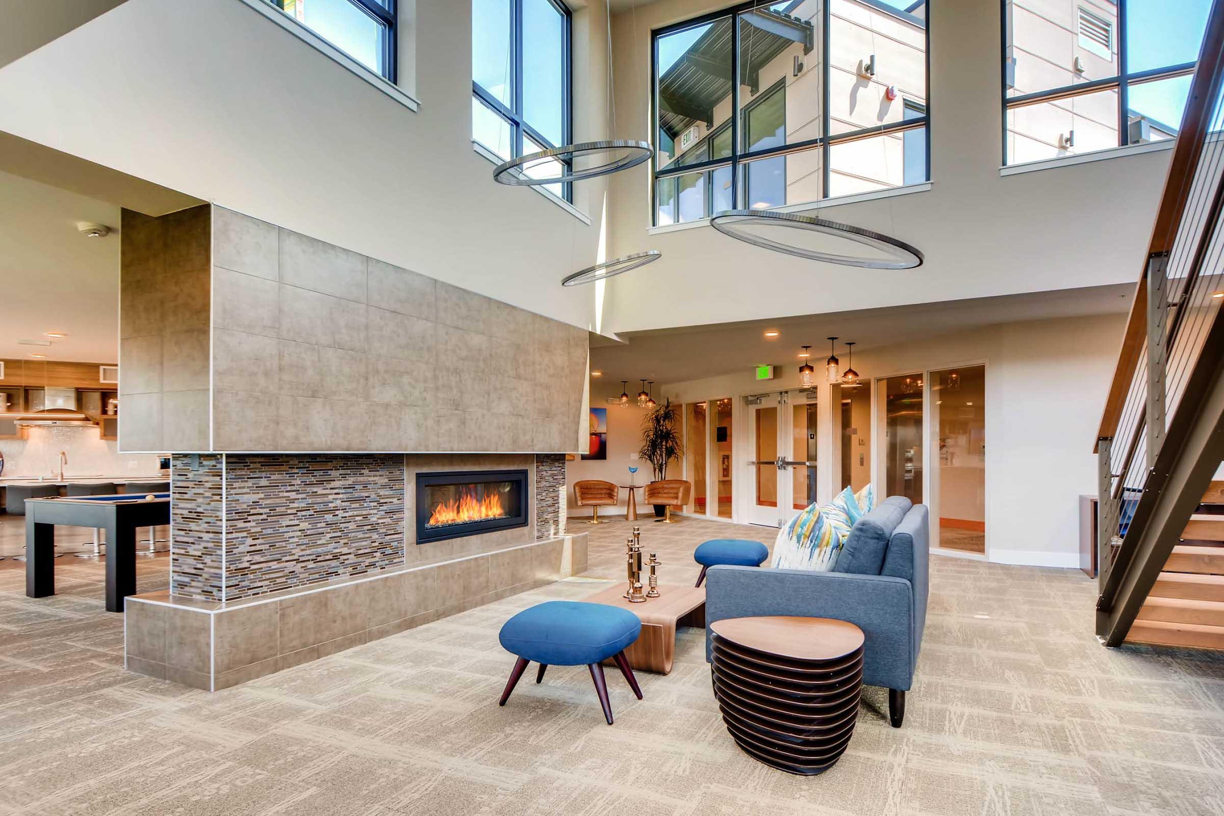 A modern living space featuring a cozy lounge area with a fireplace, large windows, and stylish furnishings. The room includes a gray sofa, circular coffee tables, and decorative accents, bathed in natural light. A staircase leads to an upper level, enhancing the contemporary design of the interior.