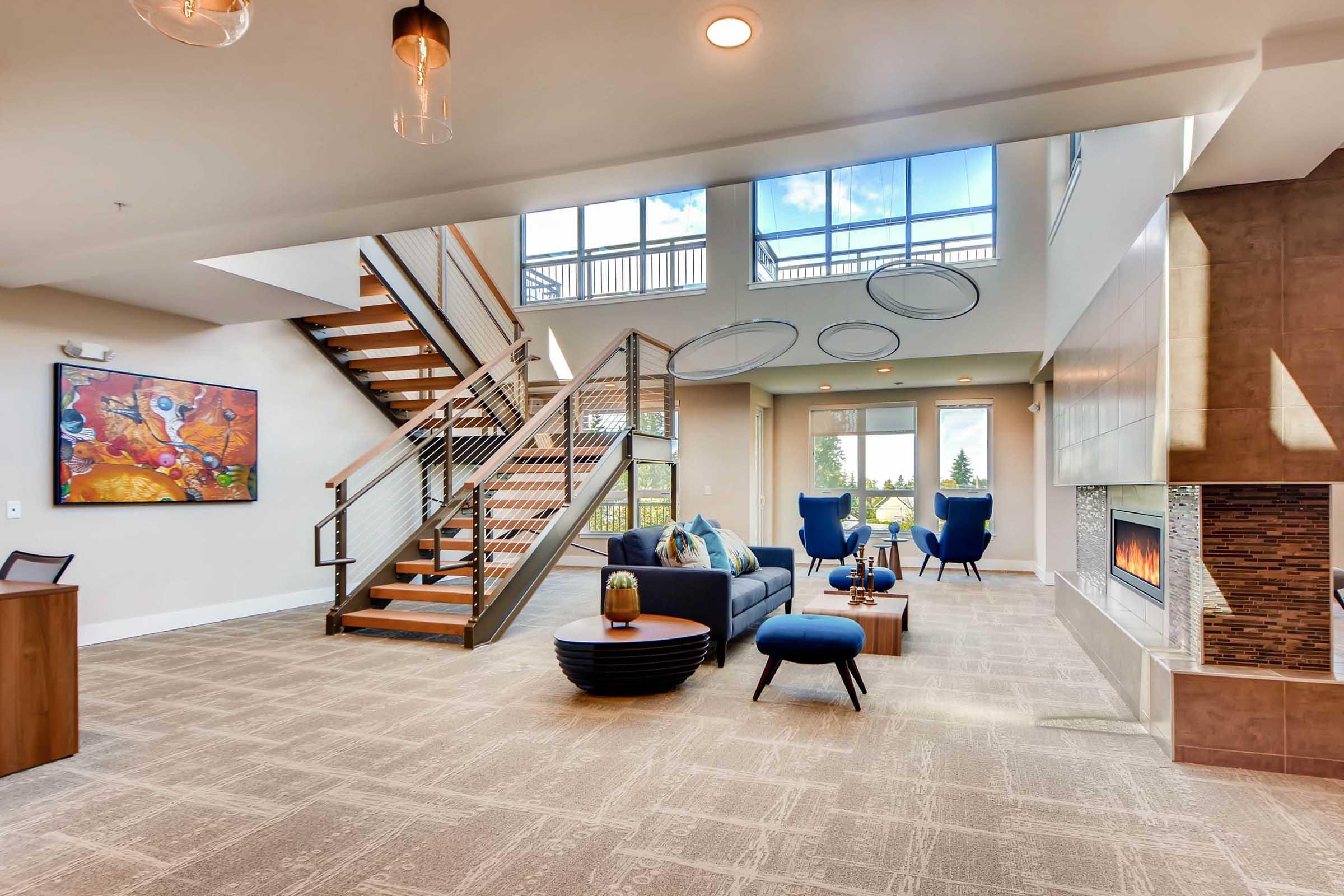 Modern interior of a spacious living area featuring a staircase, large windows, and a cozy seating arrangement with blue chairs and a dark sofa. A coffee table and decorative elements complement the warm atmosphere, while a fireplace adds a touch of elegance. Artwork decorates the walls, enhancing the inviting ambiance.
