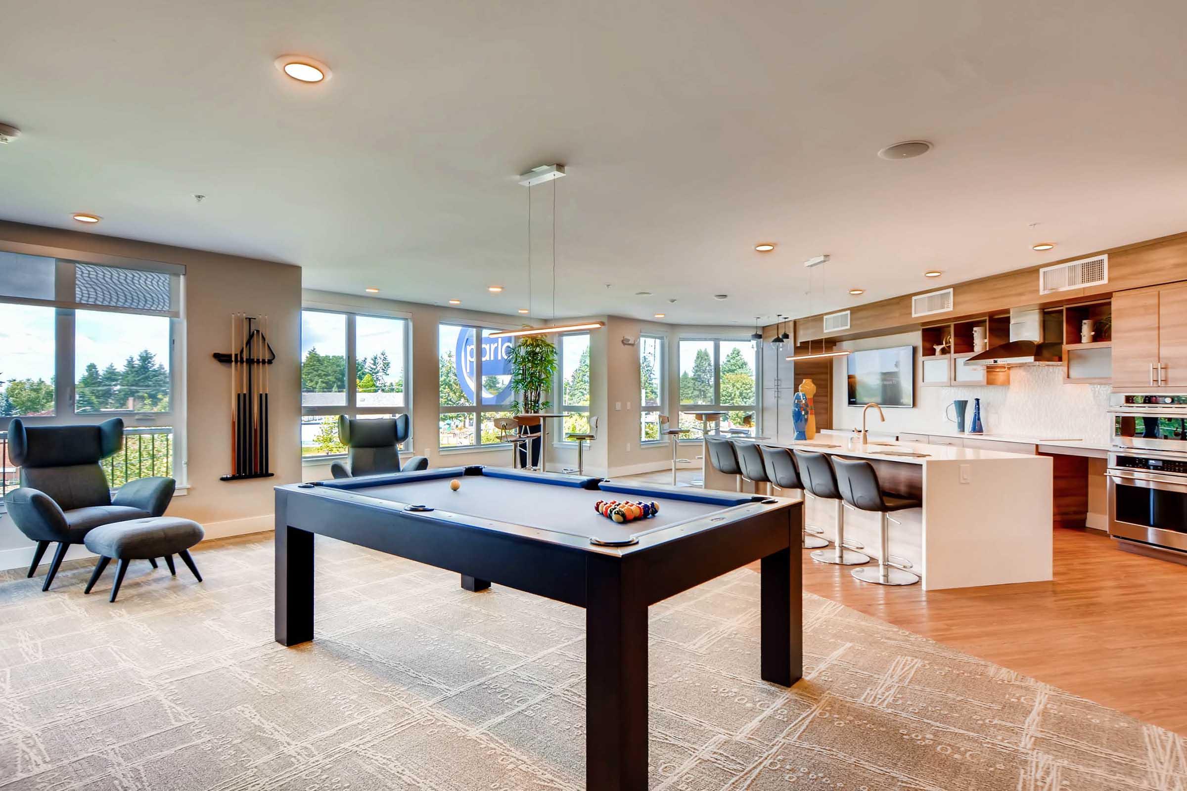 A modern interior space featuring a pool table in the foreground, with a stylish kitchen and dining area in the background. Large windows provide natural light and views of the outdoors. The decor includes contemporary furniture, including a lounge chair and bar stools.