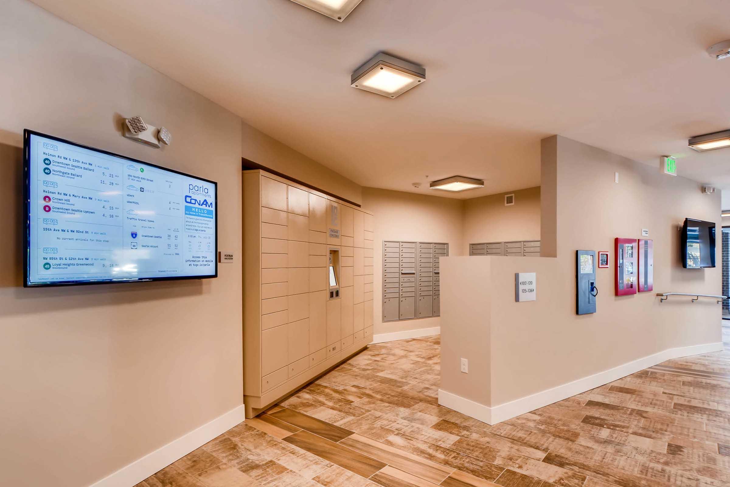 Interior view of a hallway featuring electronic screens displaying information, a row of secured mail lockers, and a small kiosk. The walls are painted in neutral tones, with modern lighting fixtures and wooden flooring.