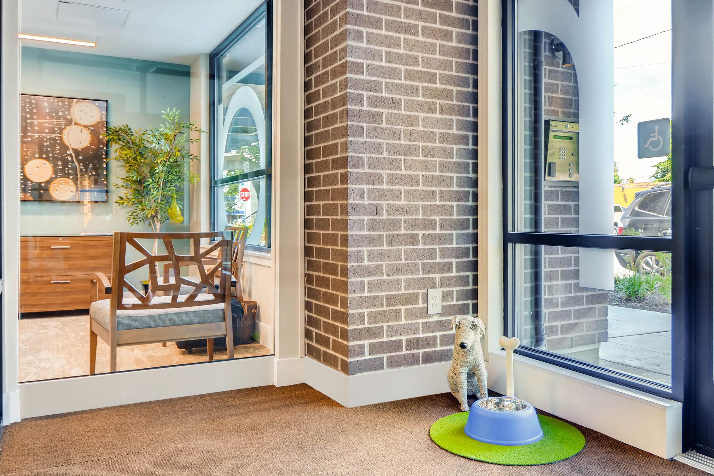 A corner of a well-lit room featuring a small dog statue next to a dog bowl on a green mat. There is a glass wall separating this area from another room, which has modern furniture and decorative plants. The floor is carpeted, and the overall ambiance is inviting and friendly.