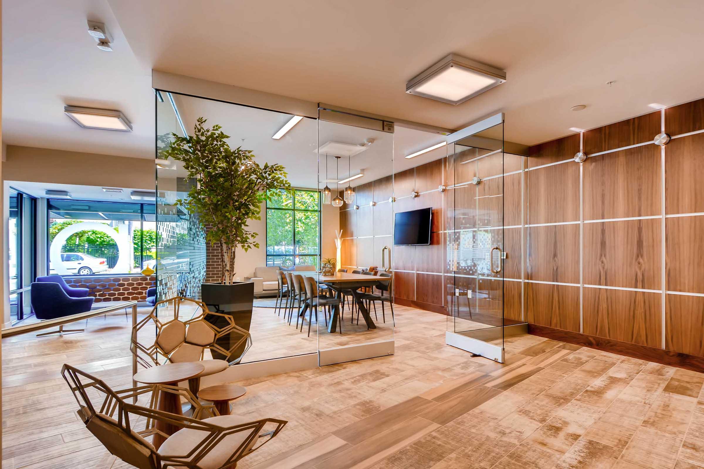 Modern office interior featuring a glass-walled conference room with a wooden accent wall, stylish furniture, a large whiteboard, and a dining table. Natural light floods the space through large windows, complemented by indoor plants and contemporary decor elements.