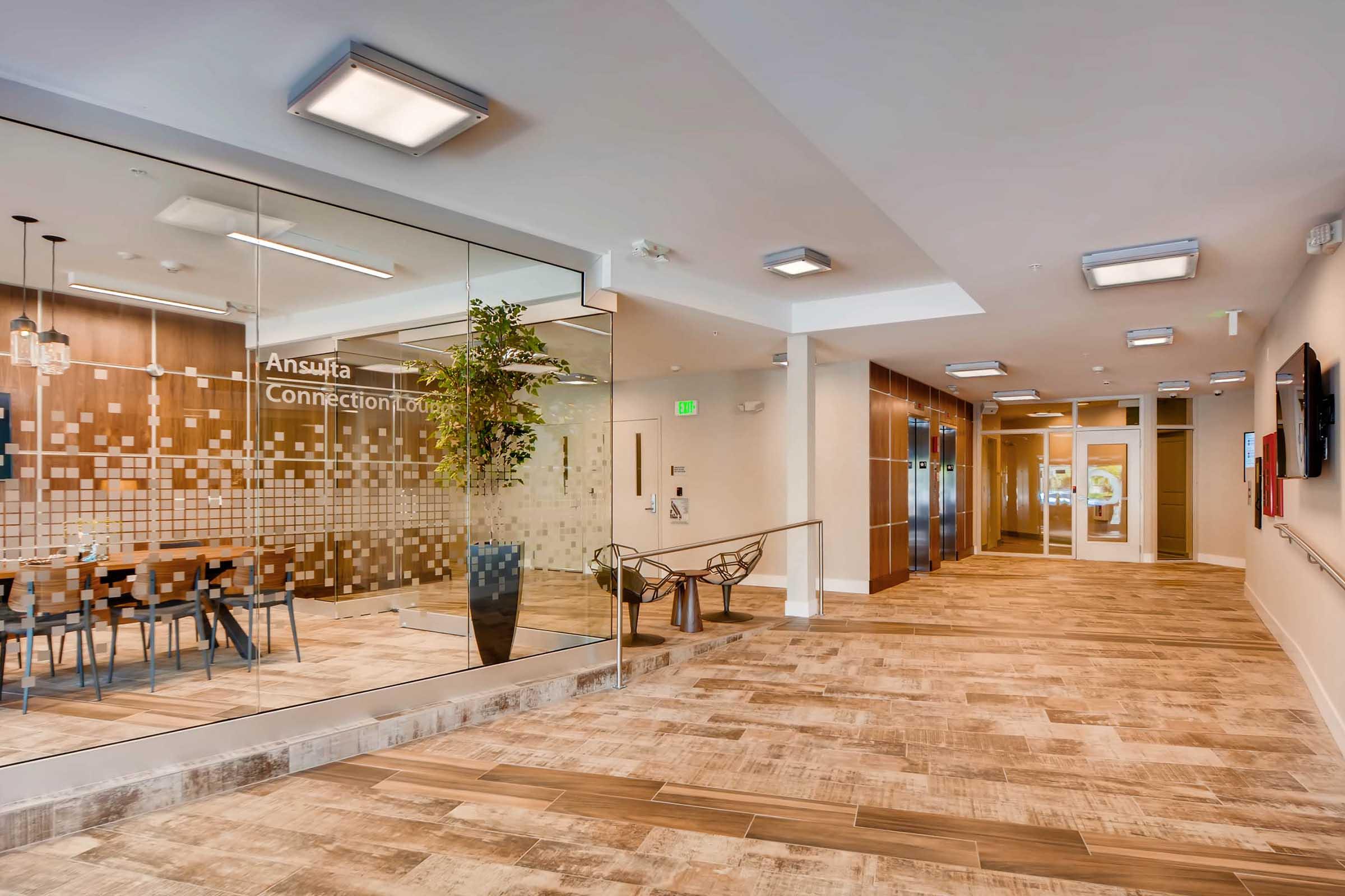 A modern hallway leading to meeting rooms, featuring a glass wall that displays the "Connection Lounge" signage. The area includes a stylish conference table with chairs and contemporary lighting, complemented by decorative plants. The flooring is designed with light-colored tiles.