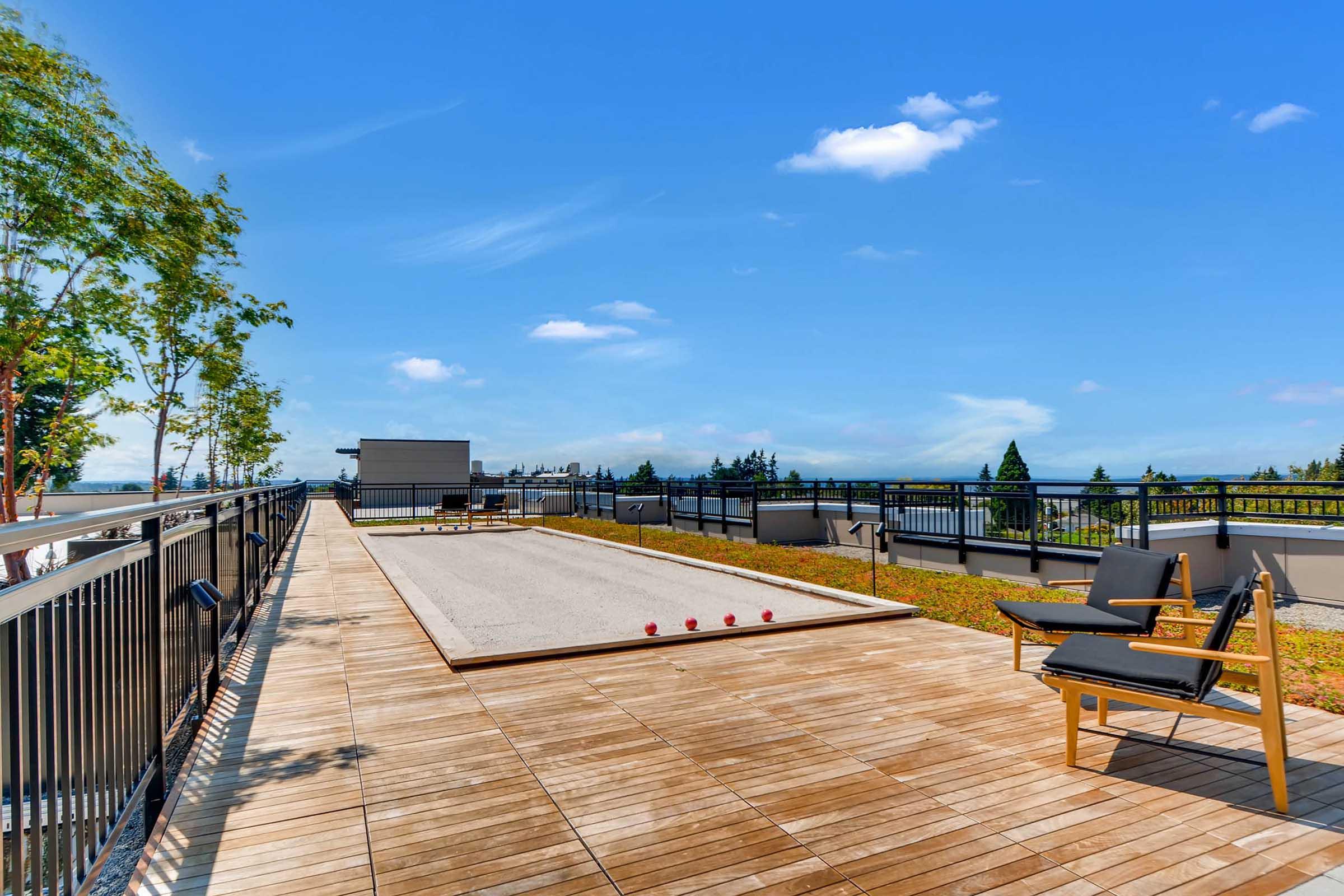 A spacious rooftop patio featuring a smooth wooden deck, a gravel bocce court with red balls, and a single modern lounge chair. Green trees line the area, framed by a clear blue sky and distant treetops, creating a relaxing outdoor space for leisure activities.