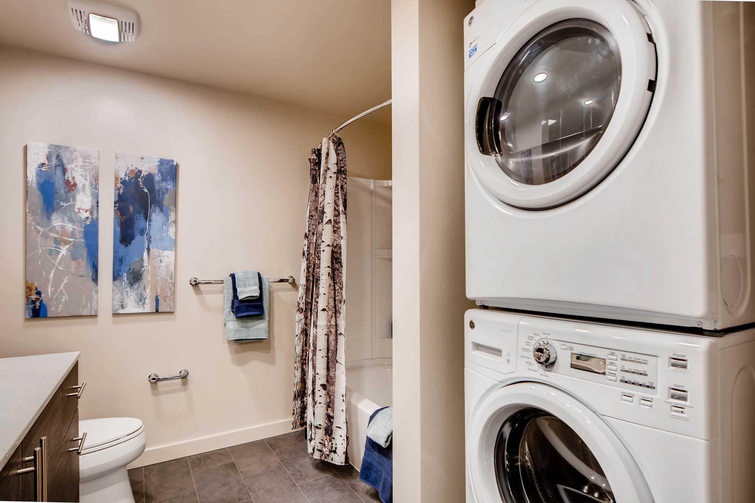 A modern bathroom featuring a stacked washer and dryer, a shower area with a curtain, and two abstract art pieces on the wall. There is a towel rack with towels and a toilet nearby, along with neutral-colored walls and tiled flooring.