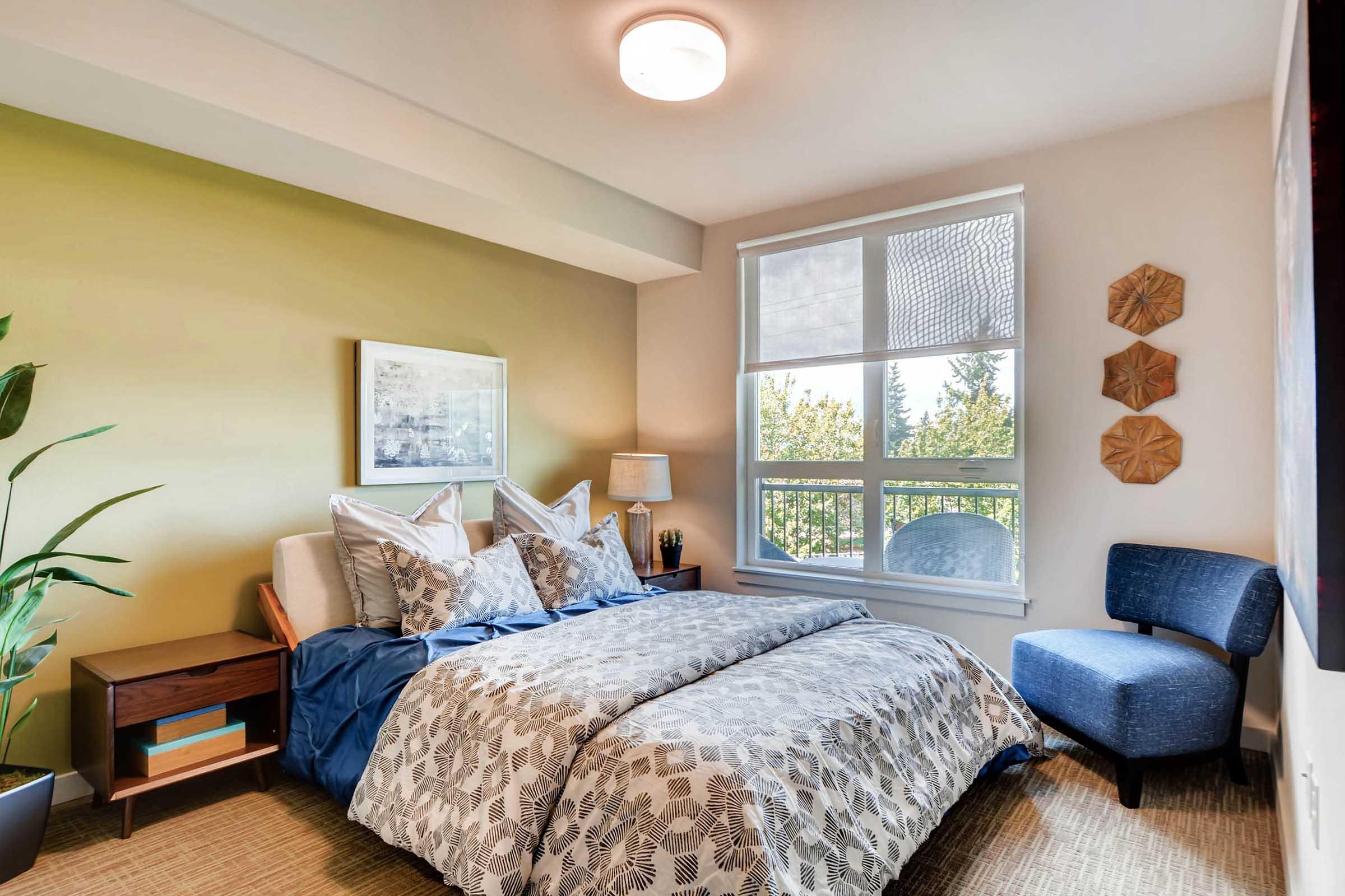 A cozy bedroom featuring a double bed with patterned bedding, two decorative pillows, and a bedside table with a lamp. A small blue armchair is positioned next to a large window, which offers a view of greenery outside. The walls are painted a soft green, creating a calming atmosphere.