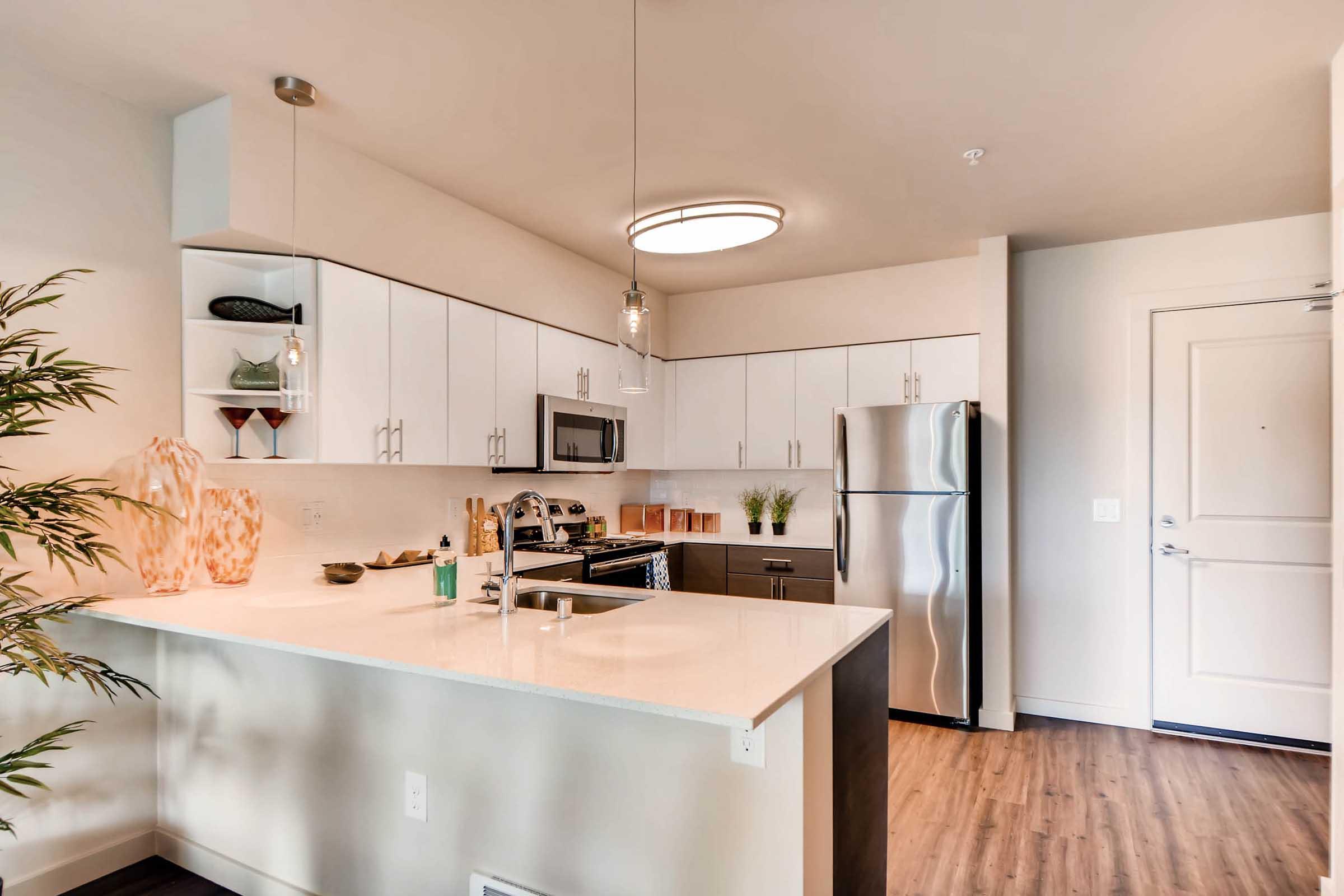Modern kitchen with white cabinetry, stainless steel appliances, and a stylish island. Features a sleek countertop, pendant lighting, and a neutral color palette. A decorative plant and glassware are displayed, enhancing the contemporary aesthetic of the space.