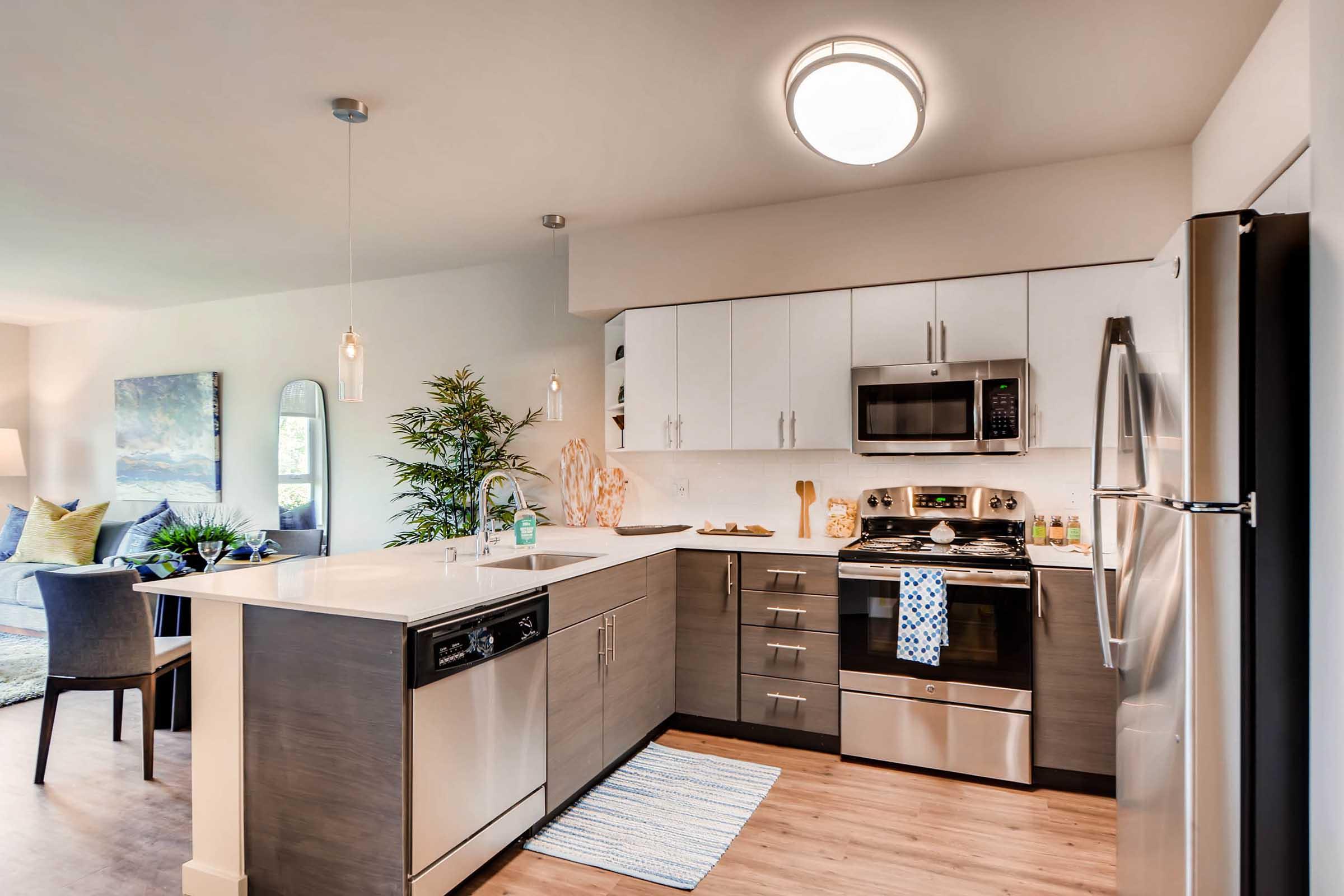 A modern kitchen featuring stainless steel appliances, including a refrigerator, oven, and microwave. Light wood flooring and cabinetry provide a contemporary look. A small island with a sink is in the center, and a dining area can be seen in the background with a comfortable chair and decorative plants. Natural light illuminates the space.