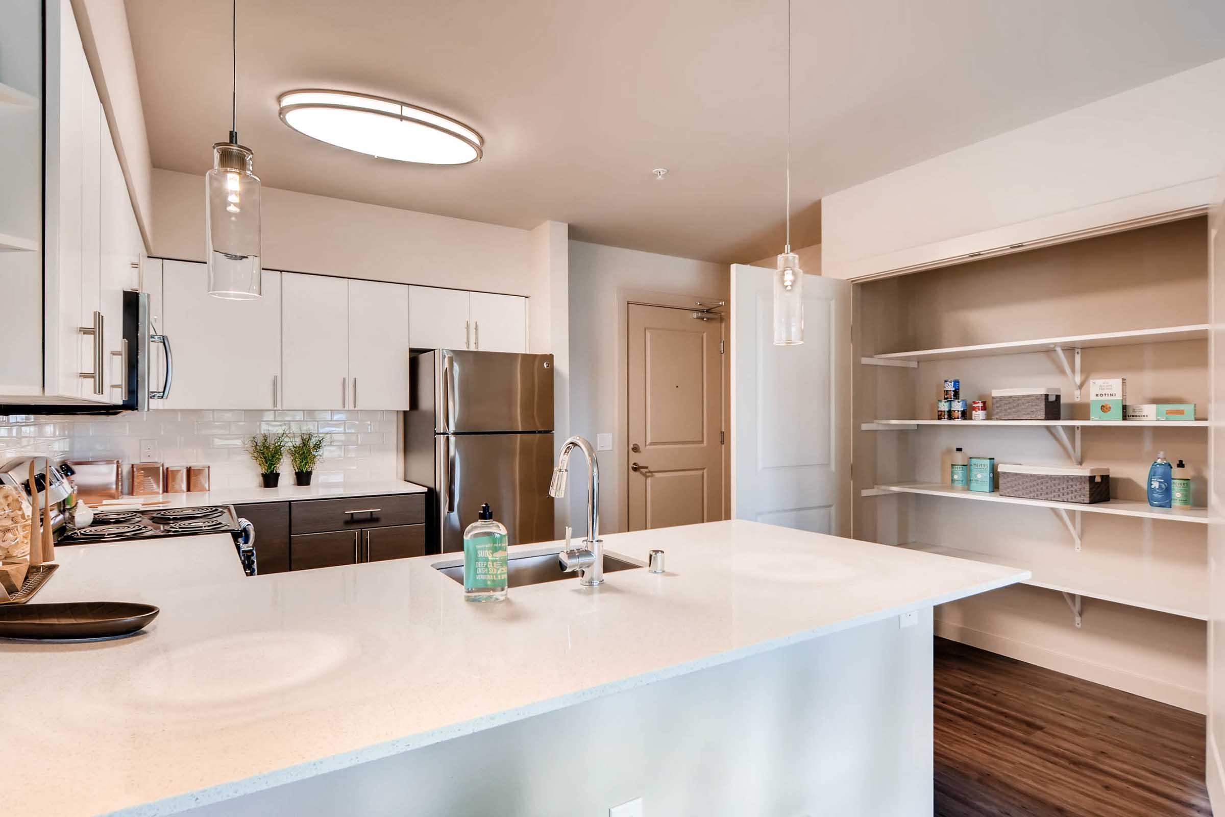 Modern kitchen with white cabinetry, a large island with a sink, stainless steel appliances including a fridge, and shelves for storage. Natural light fills the space from a ceiling fixture, creating a bright and inviting atmosphere.