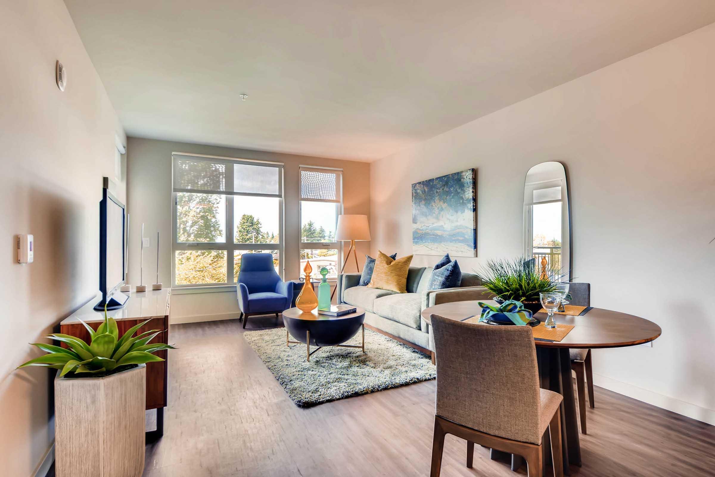 Modern living room featuring a comfortable sofa with decorative cushions, a coffee table, and a dining area with a round table and chairs. A large TV is mounted on the wall, and there are decorative plants and artwork enhancing the decor. Bright natural light enters through large windows.