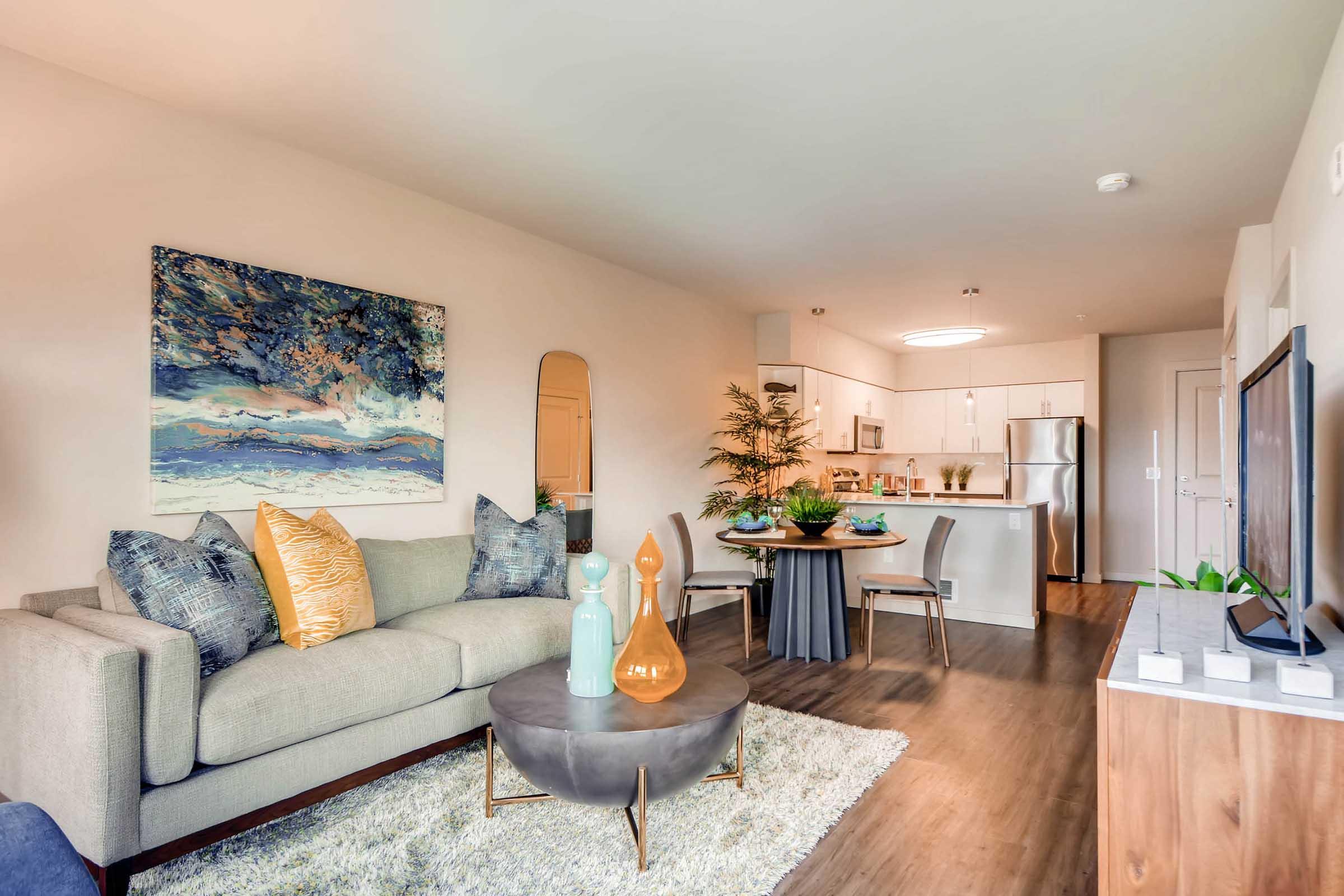 A modern living room featuring a light-colored sofa with decorative pillows, a round coffee table, and a textured rug. In the background, there's a dining area with a small table and chairs, a kitchen with stainless steel appliances, and a large artwork on the wall. The space is bright and warmly decorated with plants.