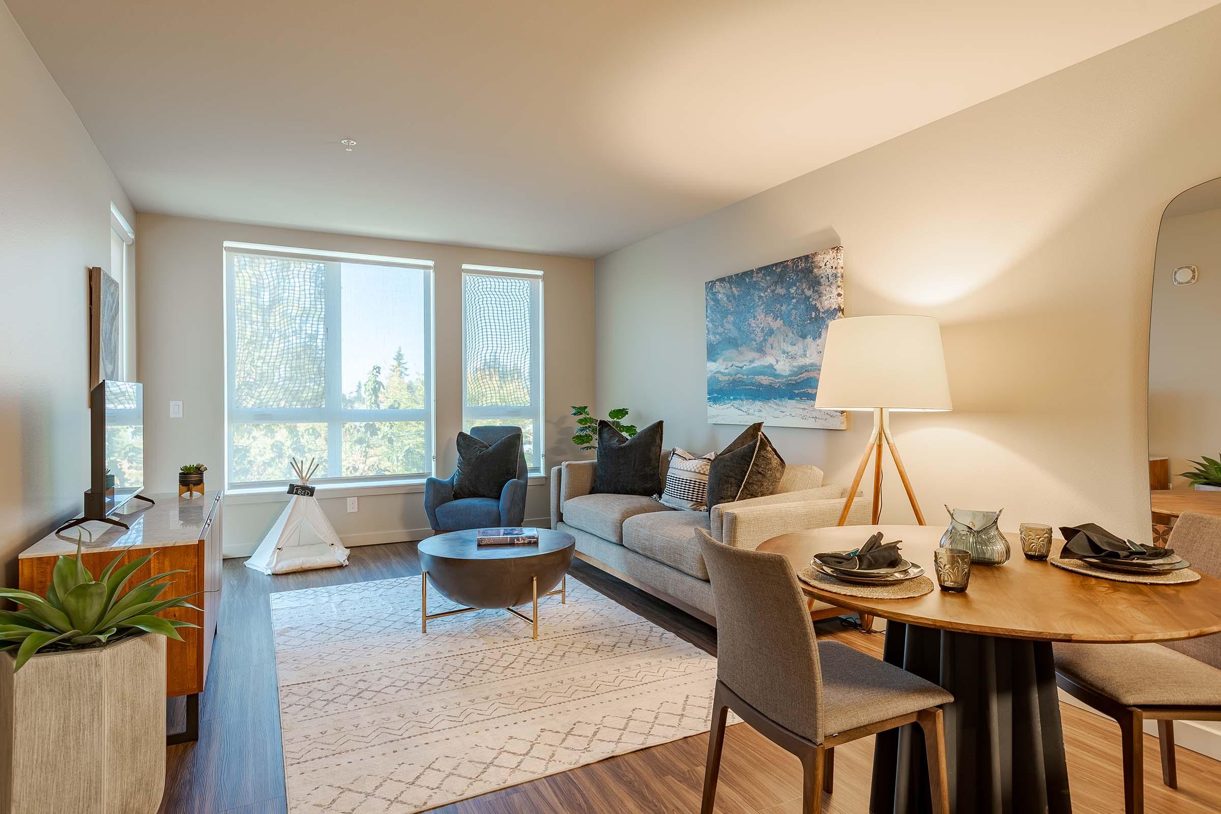 A well-decorated living room featuring a comfortable gray sofa with decorative pillows, a round coffee table, and a dining area with a small round table and chairs. Large windows allow natural light, and there are plants and artwork enhancing the cozy atmosphere. A small teepee is also visible in the corner.