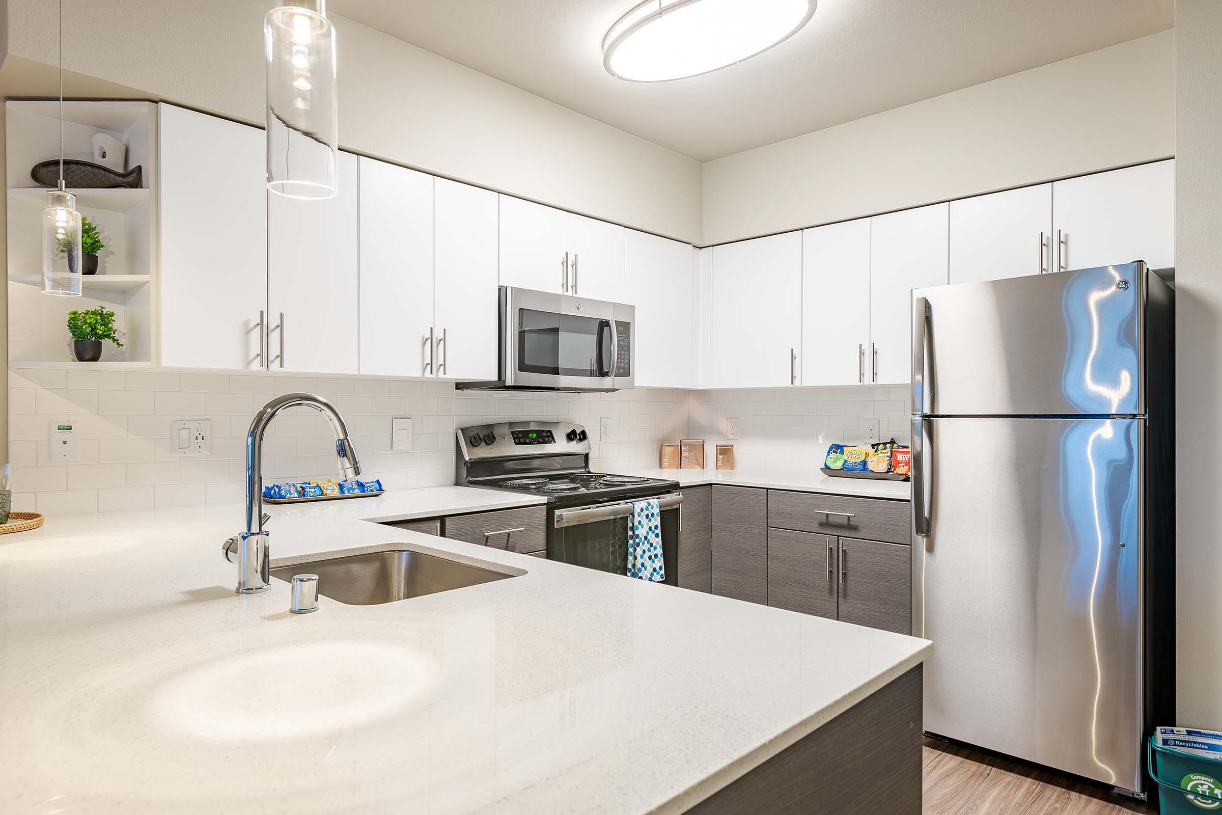 Modern kitchen with white cabinetry, stainless steel appliances including a refrigerator and microwave, a shiny countertop, and a sink with a sleek faucet. The space features pendant lighting and a small shelf with plants, creating a bright and inviting atmosphere.
