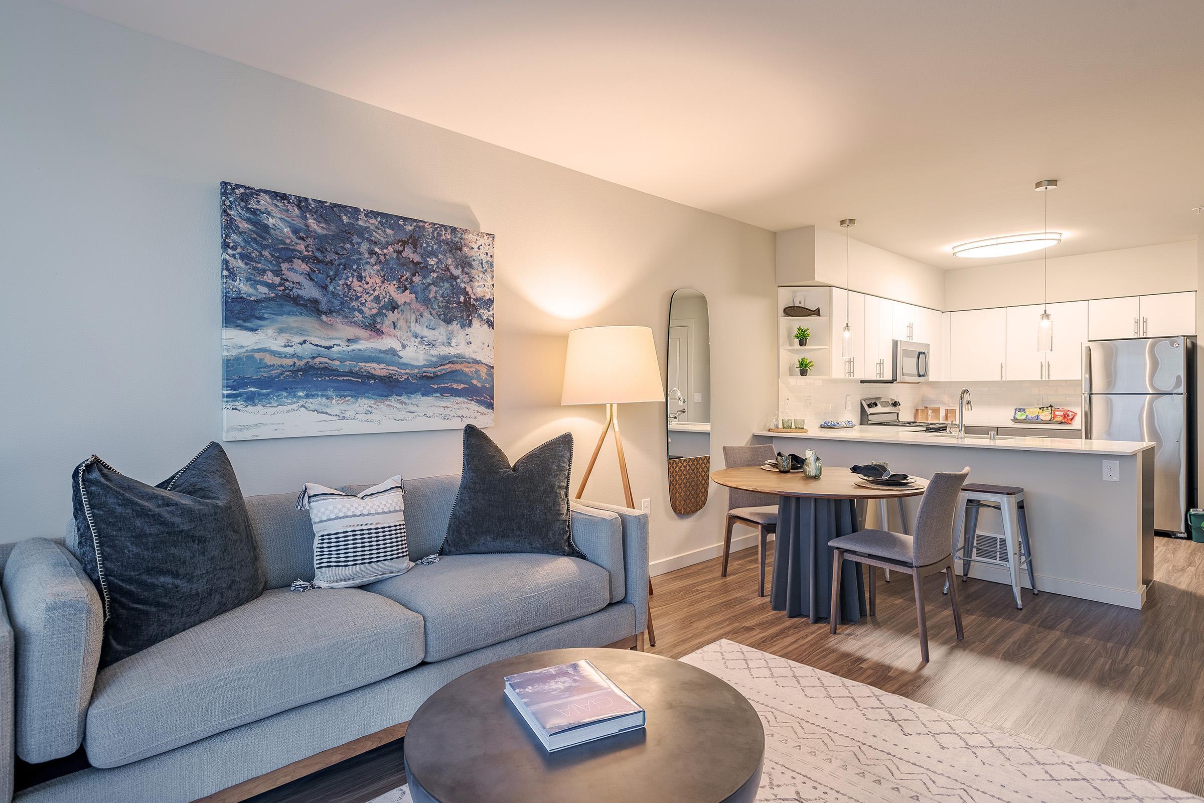 A modern living space featuring a cozy gray sofa with decorative pillows, a round coffee table with a book on it, and a dining area with a small table and chairs. The background showcases a contemporary kitchen with white cabinets, stainless steel appliances, and a piece of abstract art on the wall.