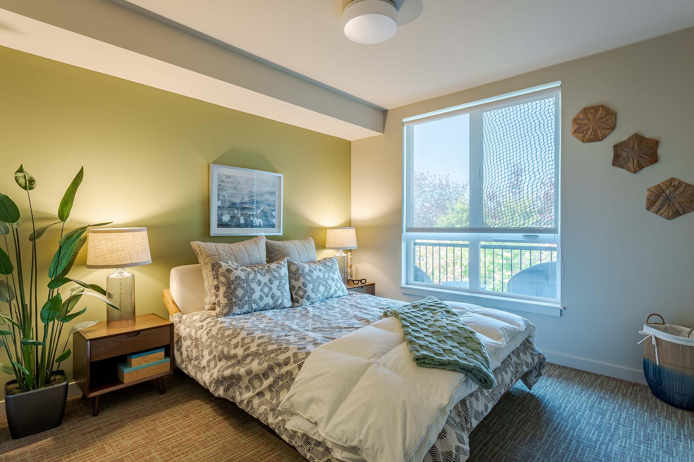 A cozy bedroom featuring a large bed with decorative pillows, two bedside lamps on wooden tables, and a green accent wall. There’s a window allowing natural light, a potted plant beside the bed, and a light-colored basket on the floor, creating a warm and inviting atmosphere.