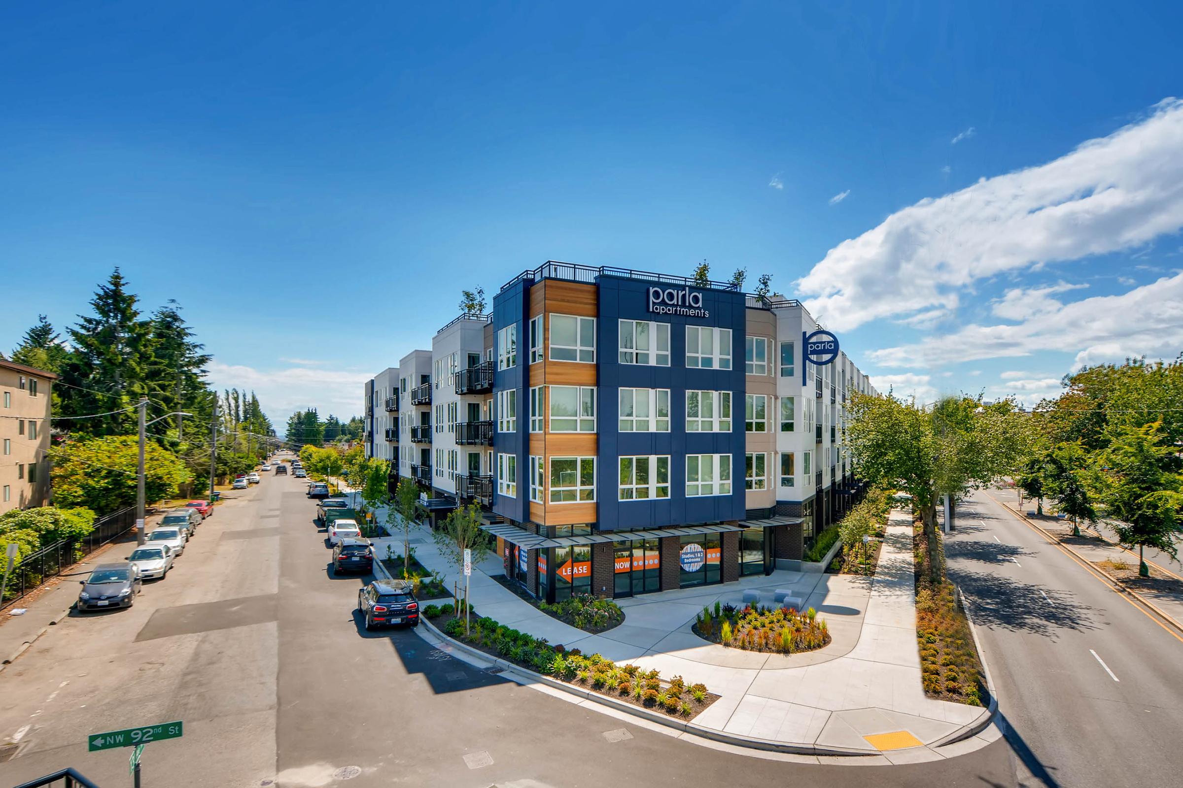 Modern apartment building at the corner of a street, featuring a mix of white and dark blue exterior with wooden accents. The scene is sunny with clear blue skies and a few fluffy clouds. There are cars parked along the street and landscaped greenery around the building.