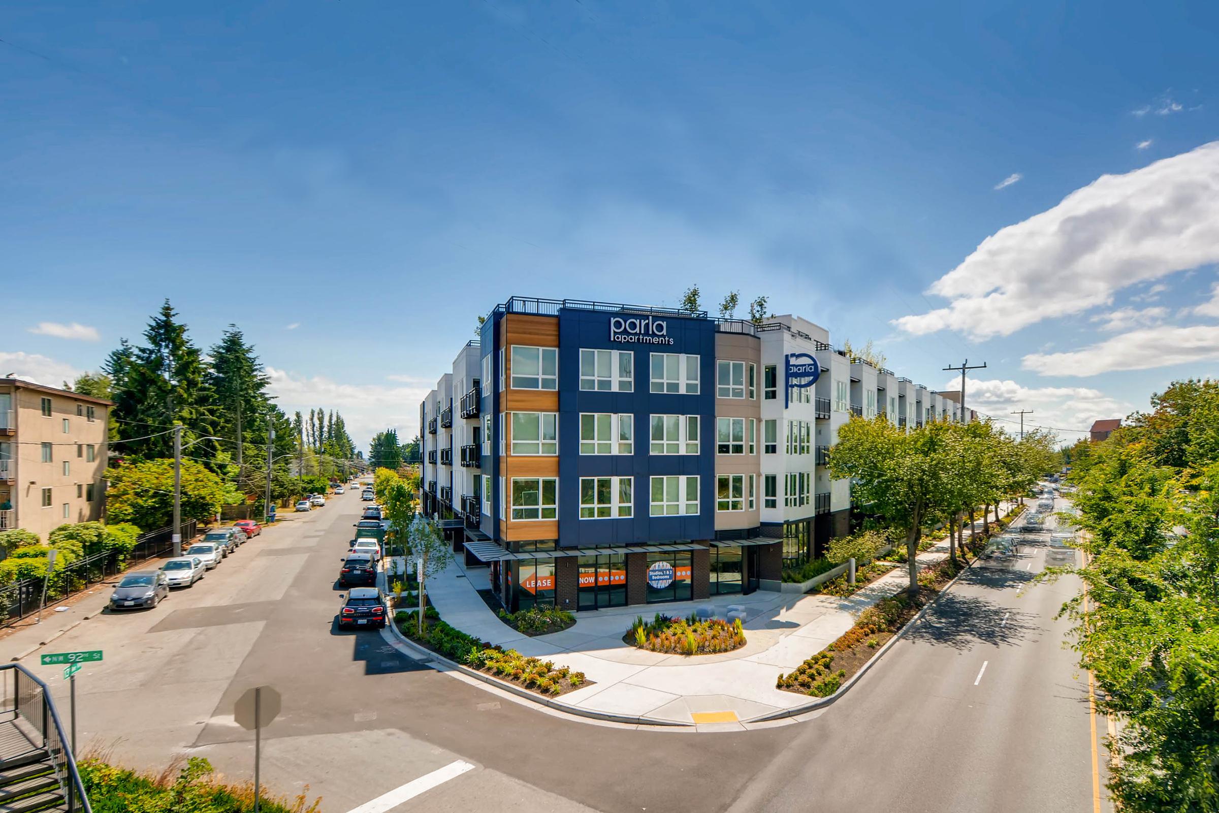 A modern apartment building with large windows and a contemporary design, located at the intersection of a tree-lined street. The building features blue and gray hues, landscaped areas, and a clear sky above with a few scattered clouds.