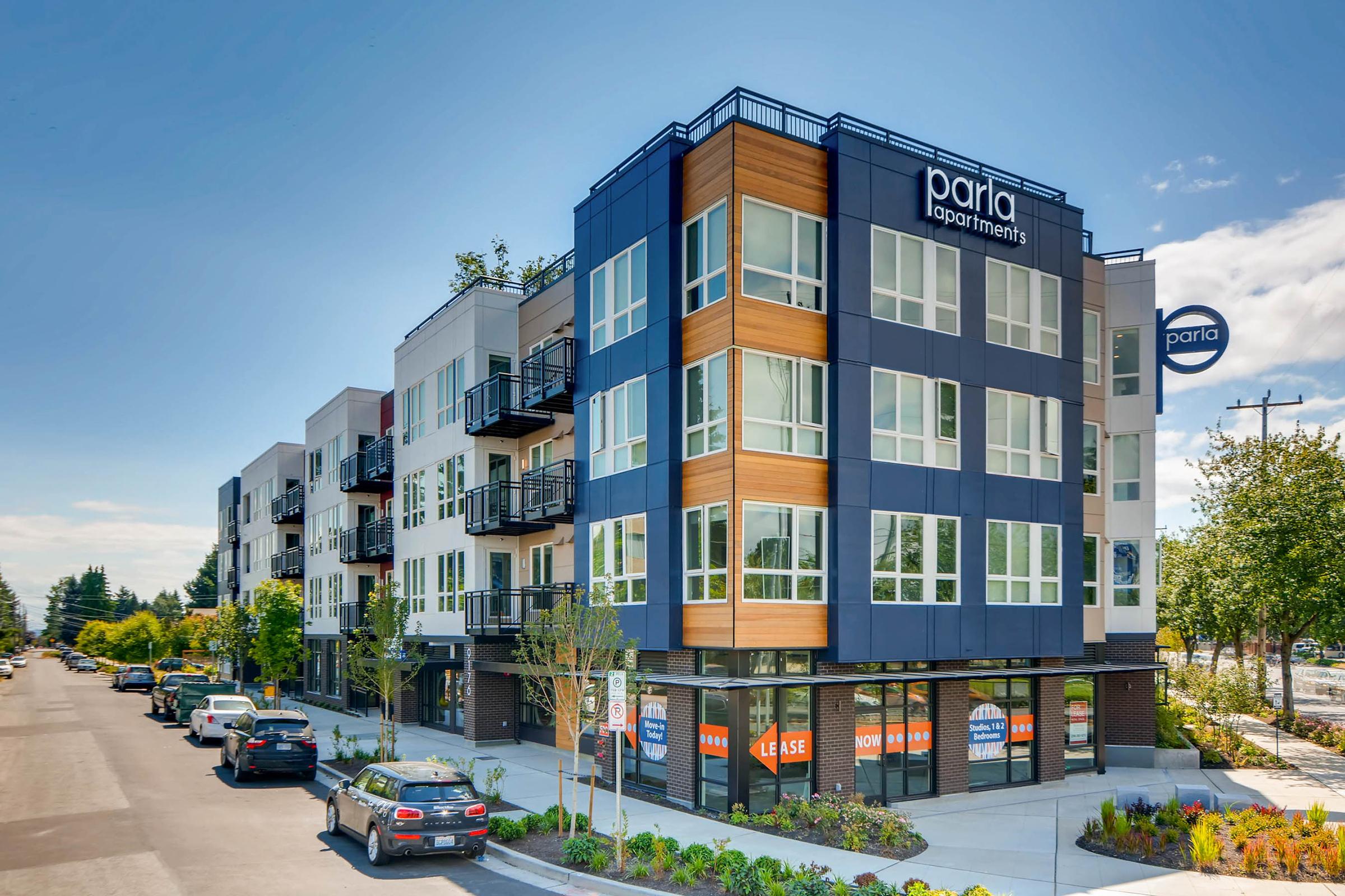 Modern apartment building with a mix of blue, white, and wood accents. The structure features large windows and balconies. The facade displays the name "Parla Apartments" prominently. Trees line the street, and parked cars are visible in front of the building. Clear blue sky enhances the scene.